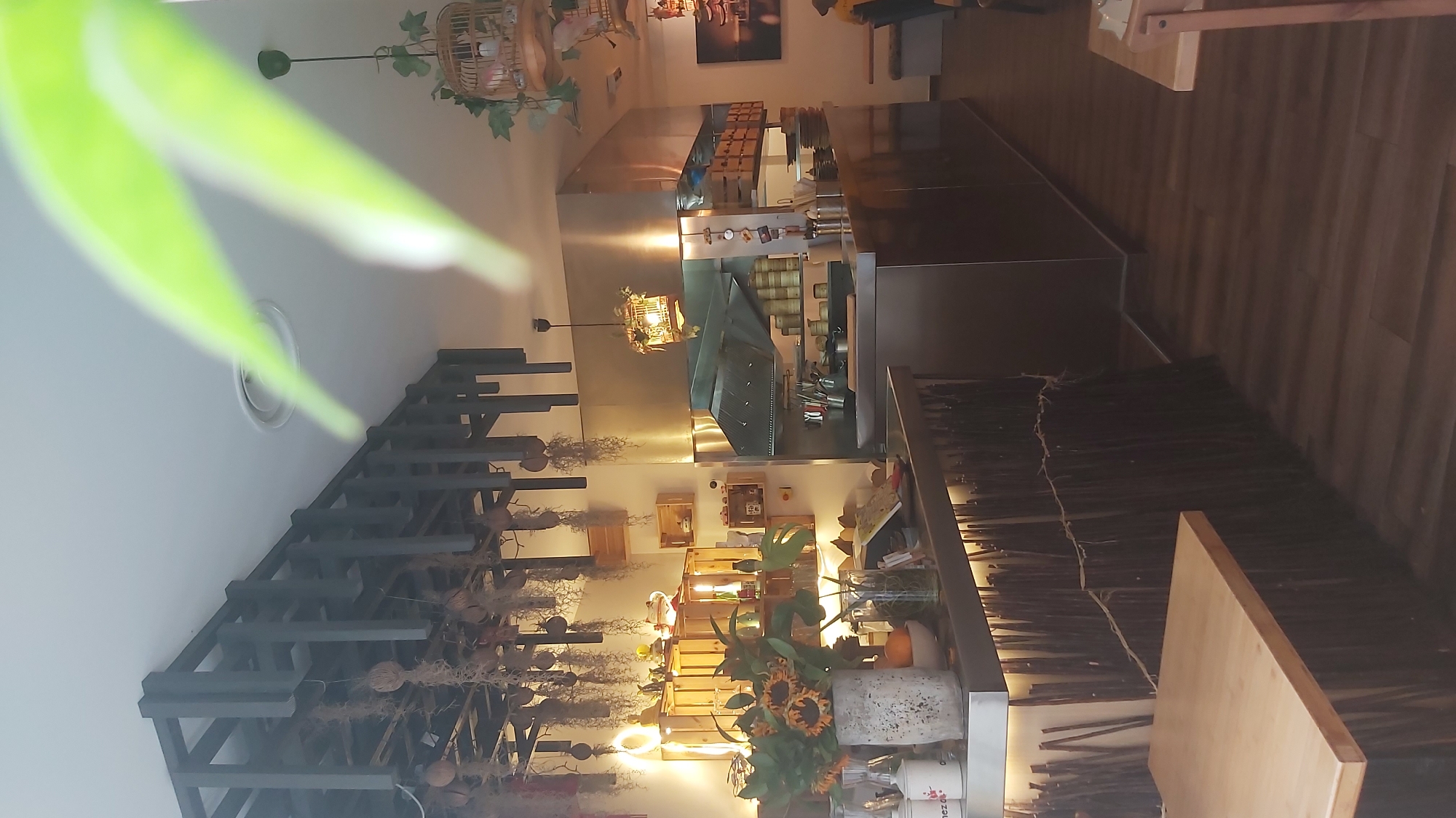 A wide-angle shot captures the interior of a restaurant, focusing on the counter and kitchen area. The decor is eclectic, with a modern industrial feel contrasted by natural elements. A black metal grate structure hangs from the ceiling, adorned with dried plants and suspended wooden boxes. A large stainless steel kitchen hood dominates the center of the frame, with various utensils and containers arranged on shelves above and around it.
To the left of the counter, a concrete vase holds a bouquet of sunflowers and greenery. Behind it, wooden shelves display decorative items, including a Buddha statue and small figures. The counter itself is made of dark wood with a rough, twig-like facade, illuminated by warm LED lights.
The flooring is dark wood laminate. In the background, a framed picture of a pagoda hangs on the wall. A small section of a dining table and a chair are visible on the right side of the image. The overall impression is of a stylish, possibly Asian-inspired, eatery.