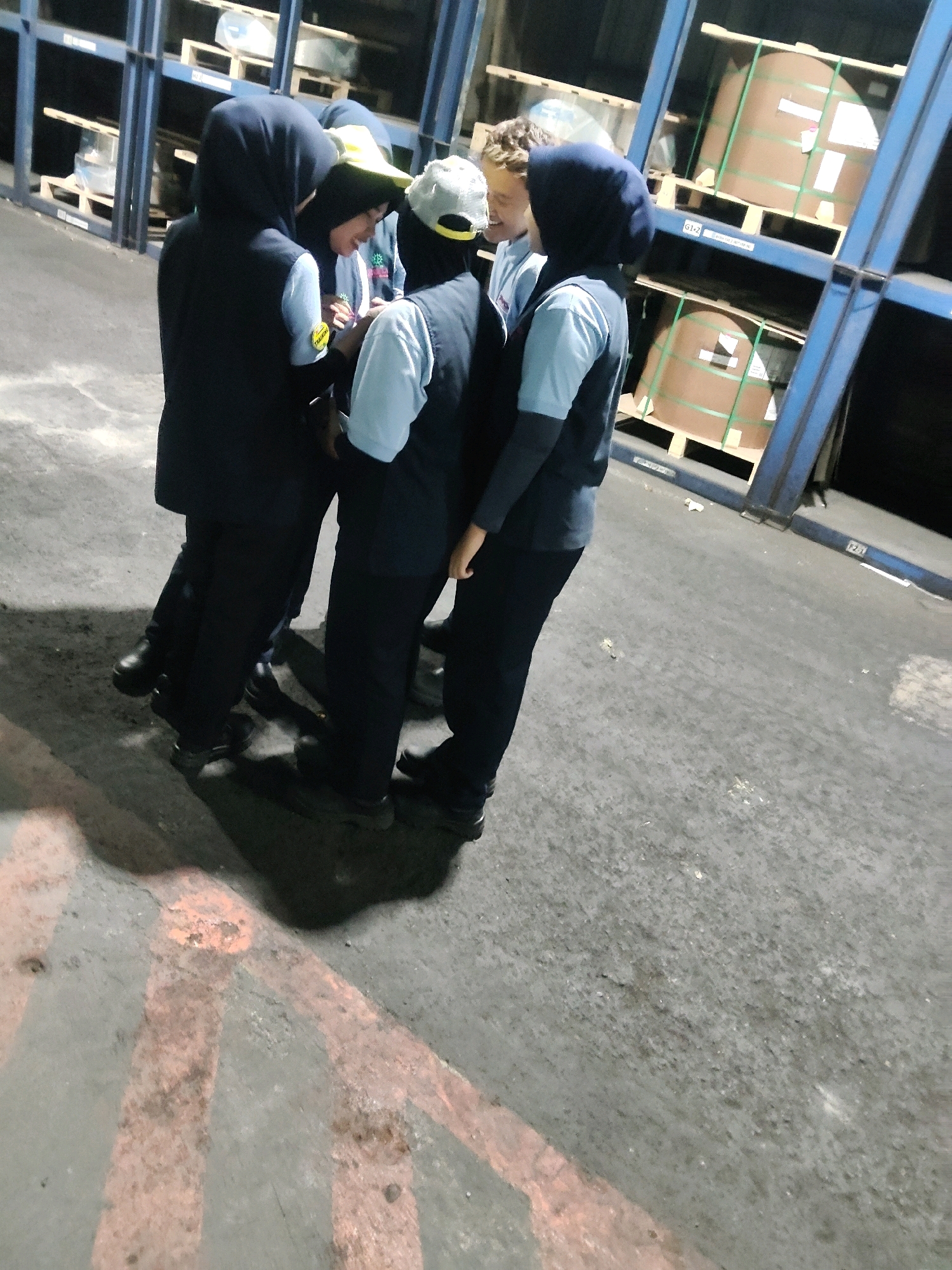 In a dimly lit indoor industrial space, possibly a warehouse or factory floor, four individuals are gathered in close proximity, their bodies partially obscured by shadows and each other. Three of the individuals appear to be women wearing dark vests over light-colored shirts and dark pants, with headscarves covering their hair. One of these women is wearing a light-colored baseball cap over her headscarf. The fourth individual, positioned between two of the women, is a person with light-colored curly hair, wearing a light-colored shirt. They are all facing inwards, looking down at something held by one of the women, suggesting a collaborative or shared activity.
The environment is characterized by a concrete floor with faded orange markings, possibly indicating pathways or designated areas. In the background, tall blue metal shelving units are visible, stacked with large, cylindrical objects wrapped in brown paper and secured with green strapping, resembling rolls of material such as paper, fabric, or metal. The lighting is artificial, casting long shadows and creating a somewhat grainy texture to the image. The overall scene conveys a sense of work or a brief pause in activity within an operational facility. The mention of Mekarmukti, Indonesia, suggests this location is within that region.
