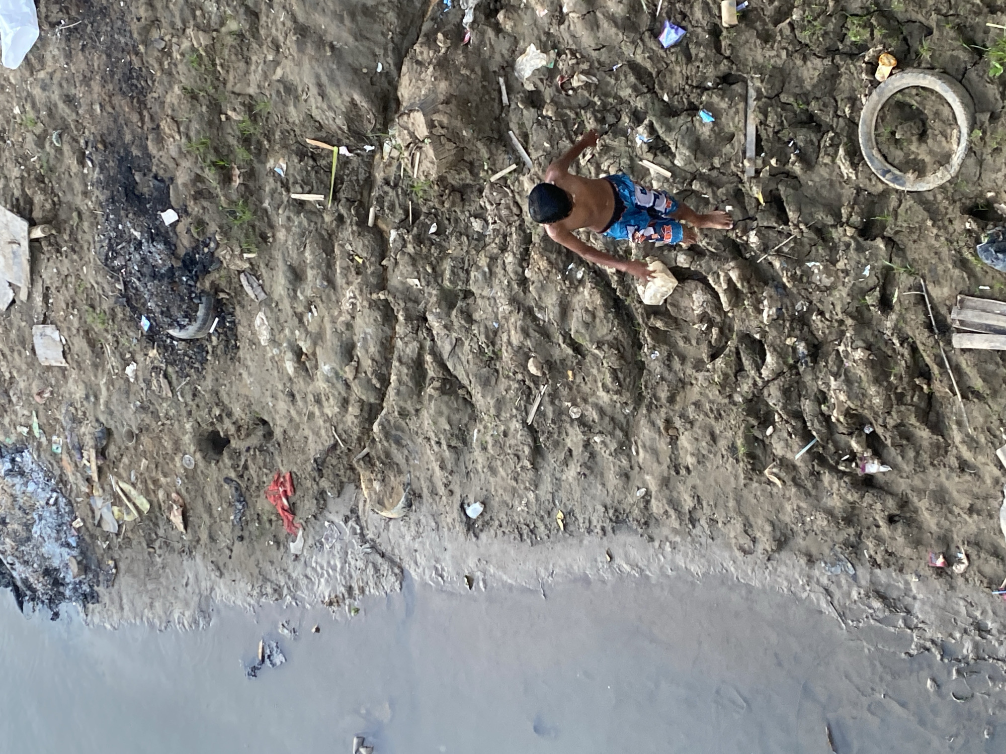 An aerial view captures a boy in blue and white patterned shorts walking on a muddy, debris-strewn embankment beside a body of water. The embankment is littered with trash, including plastic bags, shredded material, and a discarded tire. The water appears murky and still. The scene is consistent with the bank of the Musi River in Tunggul Irang Ulu, Indonesia, a region known for its informal settlements and associated waste management challenges.