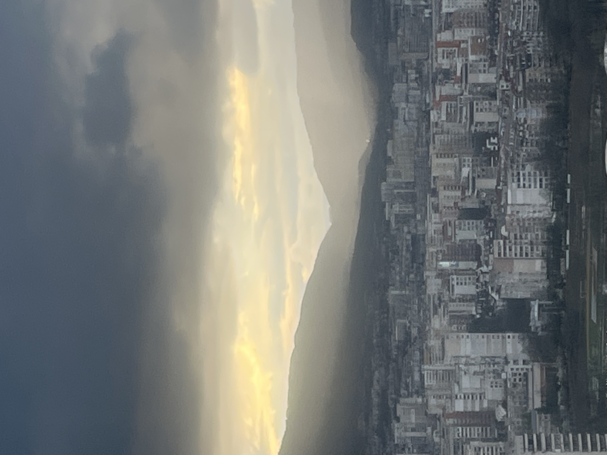 The image shows a view of Sofia, Bulgaria, with a mixture of dark rain clouds, bright sunlight, and a mountain range. On the left side of the image are dark storm clouds. There is a line of sunlight illuminating the edge of the mountains in the middle of the frame. The right side depicts the urban landscape of Sofia with its buildings, streets, and trees.