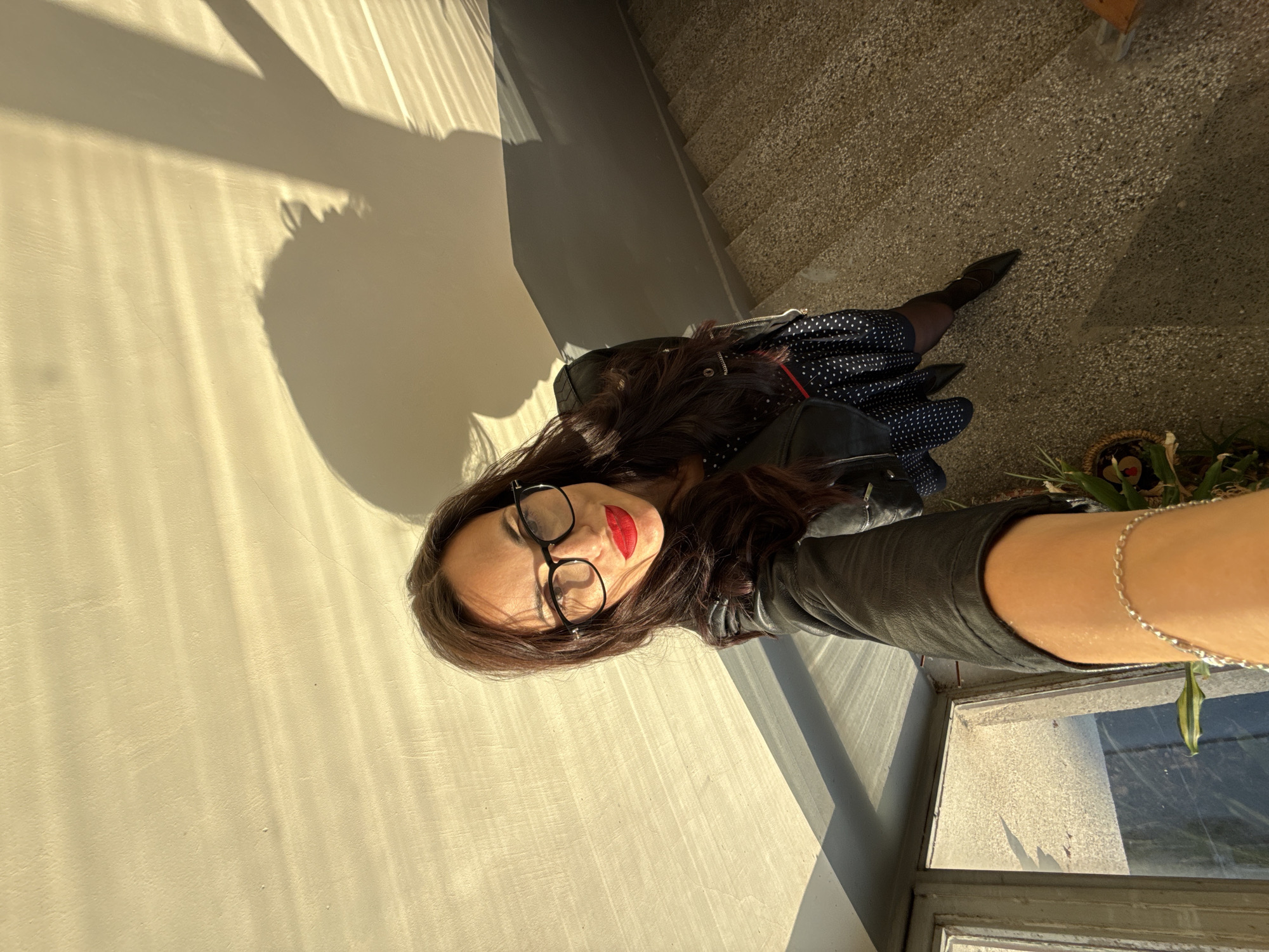 A woman with dark hair and glasses, wearing a black leather jacket and a blue polka-dot dress, is taking a selfie indoors. She is positioned near a window, with a sunlit wall to her left and stairs in the background. A plant is visible near the window. The location is Pleven, Bulgaria.