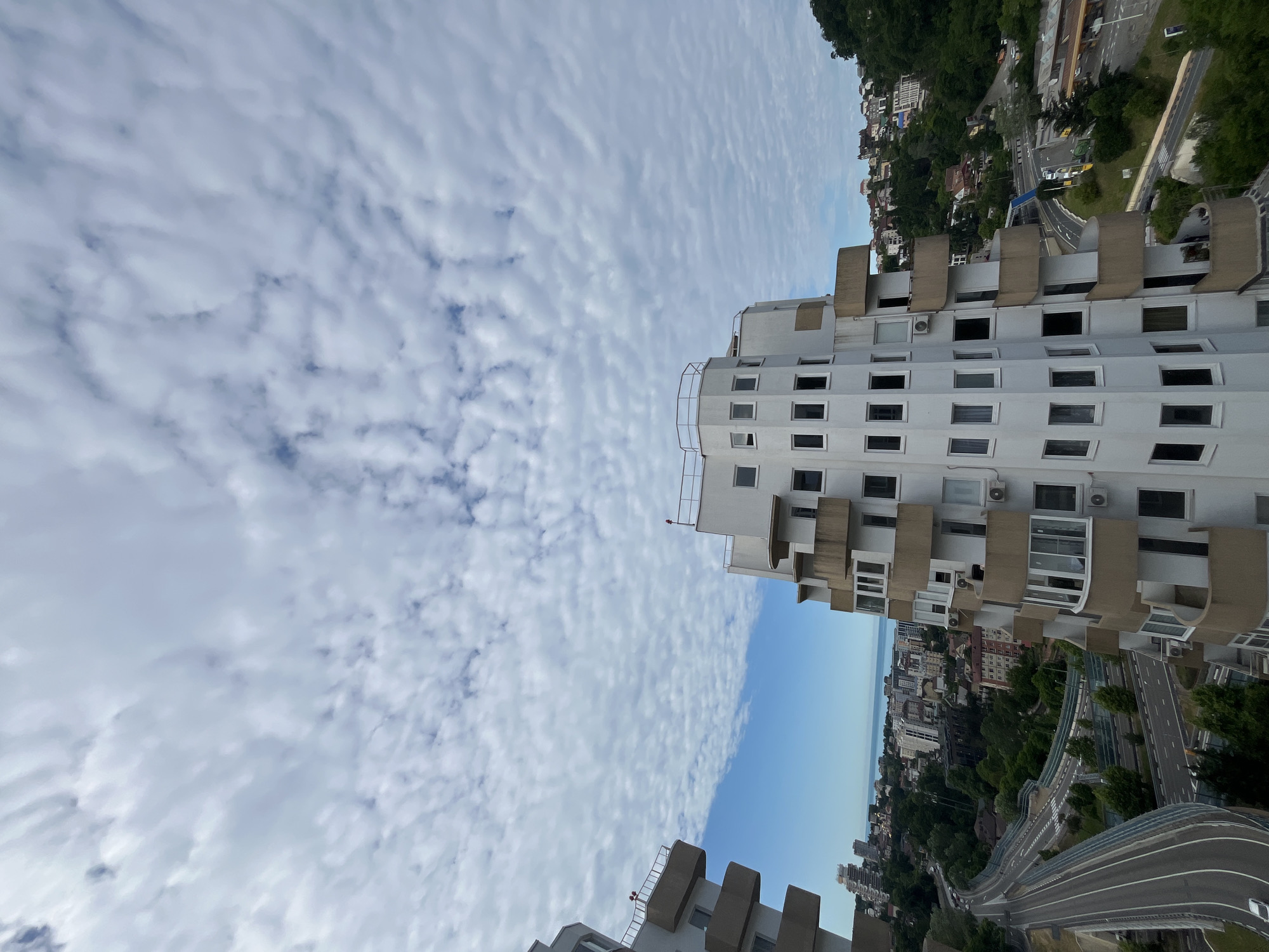 This is a high-angle, full shot of a modern white apartment building with uniquely shaped balconies, located in Sochi, Russia. The building occupies the foreground and right-middle of the frame. Its facade is a repeating pattern of windows and protruding balconies with beige trim. The balconies are curved and staggered, creating an undulating effect. The top of the building has a railing and what appears to be a small antenna.
To the left of the main building, another similar building is partially visible, also with staggered balconies. In the background, the city of Sochi sprawls out, with other apartment buildings and residential areas visible. The buildings are nestled amongst lush green trees and rolling hills. A multi-lane road curves through the landscape in the mid-ground, with traffic visible, including a white car on the left side of the road. Further in the distance, the Black Sea is visible, meeting the horizon under a sky filled with textured, mottled white clouds. The overall impression is of a modern, urban environment integrated with nature, seen from a high vantage point on a cloudy day.
