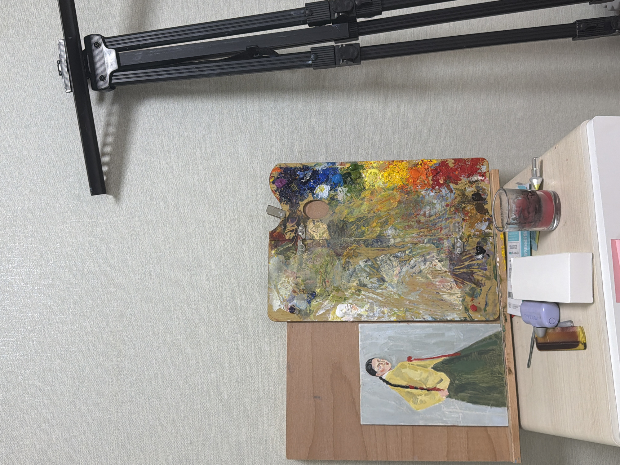 This is a still life photograph featuring art supplies and a painting, possibly in an indoor studio space. The background is a textured light-colored wall. To the right, a black tripod is partially visible, casting a shadow on the wall. In the center, a wooden artist's palette is heavily laden with dried and wet oil paints in a vibrant array of colors. To the left of the palette is a small, framed painting depicting a person in traditional Korean attire, a hanbok, with their back turned to the viewer, looking over their shoulder. The painting shows the figure wearing a yellow jacket and a green skirt, with a red sash. Beneath the painting and palette, a light-colored table or surface holds several items. These include a small glass containing a dark red substance, a tube of paint, a small white box, and a comb. The scene appears to be well-lit, possibly by artificial light, highlighting the textures and colors of the art supplies. The location is stated to be in Dongducheon-si, South Korea.