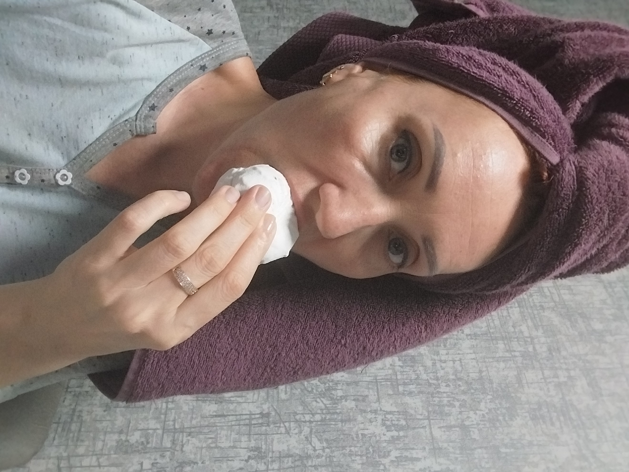 A woman with a purple towel wrapped around her head holds a white cotton pad to her mouth. She is wearing a gray and blue pajama top with a star pattern and a button-up neckline. The woman has fair skin, dark eyebrows, and visible pores and lines on her face. She is also wearing a gold band on her left ring finger. The background is a gray, textured wall. The image is a close-up shot, cropped to focus on the woman's face and upper body. There is no identifiable information within the image to determine its location in Dnipro, Ukraine, or any specific environment beyond a typical indoor setting.