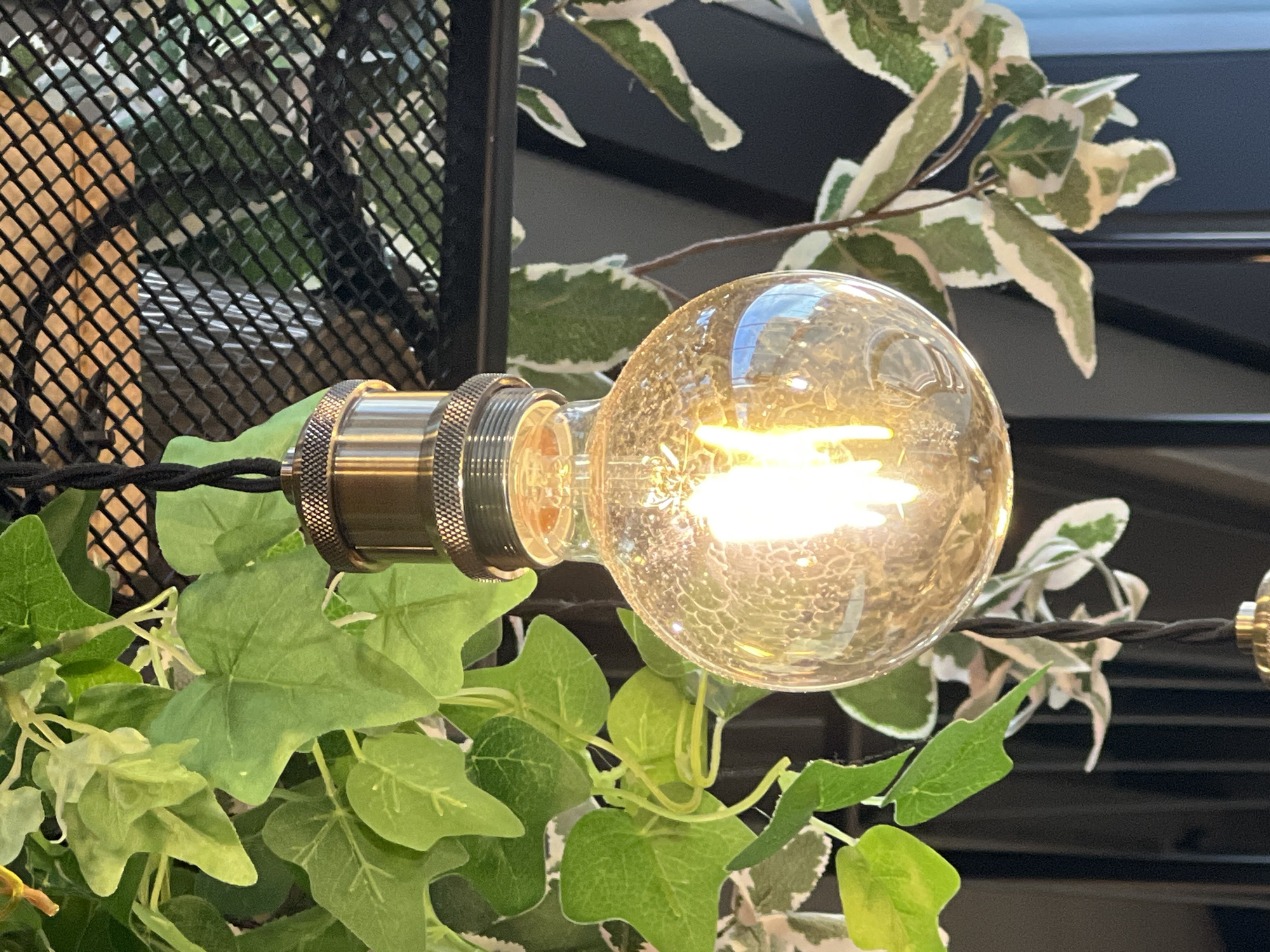 This image shows a close-up view of a vintage-style Edison light bulb hanging from a black cord. The bulb is lit, emitting a warm yellow glow. It is surrounded by lush green foliage, with some leaves appearing to be artificial. Behind the foliage, a black metal mesh grate is visible. The scene appears to be indoors, possibly in a cafe or restaurant, with a dark background. The lighting is soft and diffused. The image does not contain any people or indicate any specific location within Sinyavino, Russia.