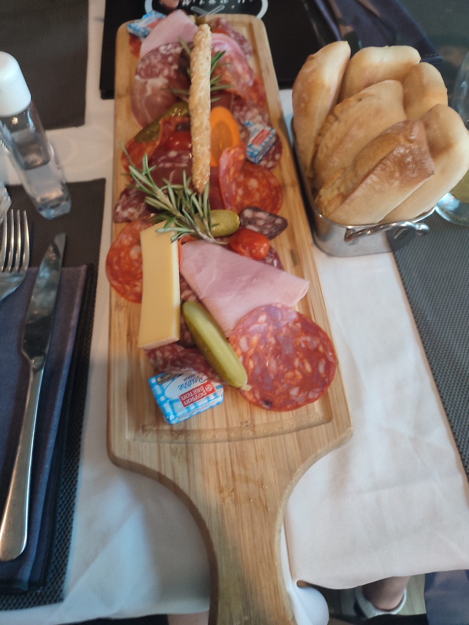 An overhead, eye-level shot shows a charcuterie board on a wooden serving platter at a table. The platter contains various cured meats, including sliced salami and ham, a cheese wedge, a whole pickle, a breadstick, and an olive. A small butter packet with blue and white checkered paper is also visible on the platter. To the left of the platter are a fork and knife resting on a dark grey napkin. A glass bottle, possibly containing water or hand sanitizer, stands to the far left of the frame. Behind the platter, a metal basket holds several pieces of bread. The table is covered with a white tablecloth. A small portion of a person's legs and feet, wearing white sneakers, are visible at the bottom right of the image. The background is blurred but suggests an indoor setting, possibly a restaurant. The lighting appears to be ambient and somewhat dim. The scene is devoid of explicit indicators of the city of Hyères, France.