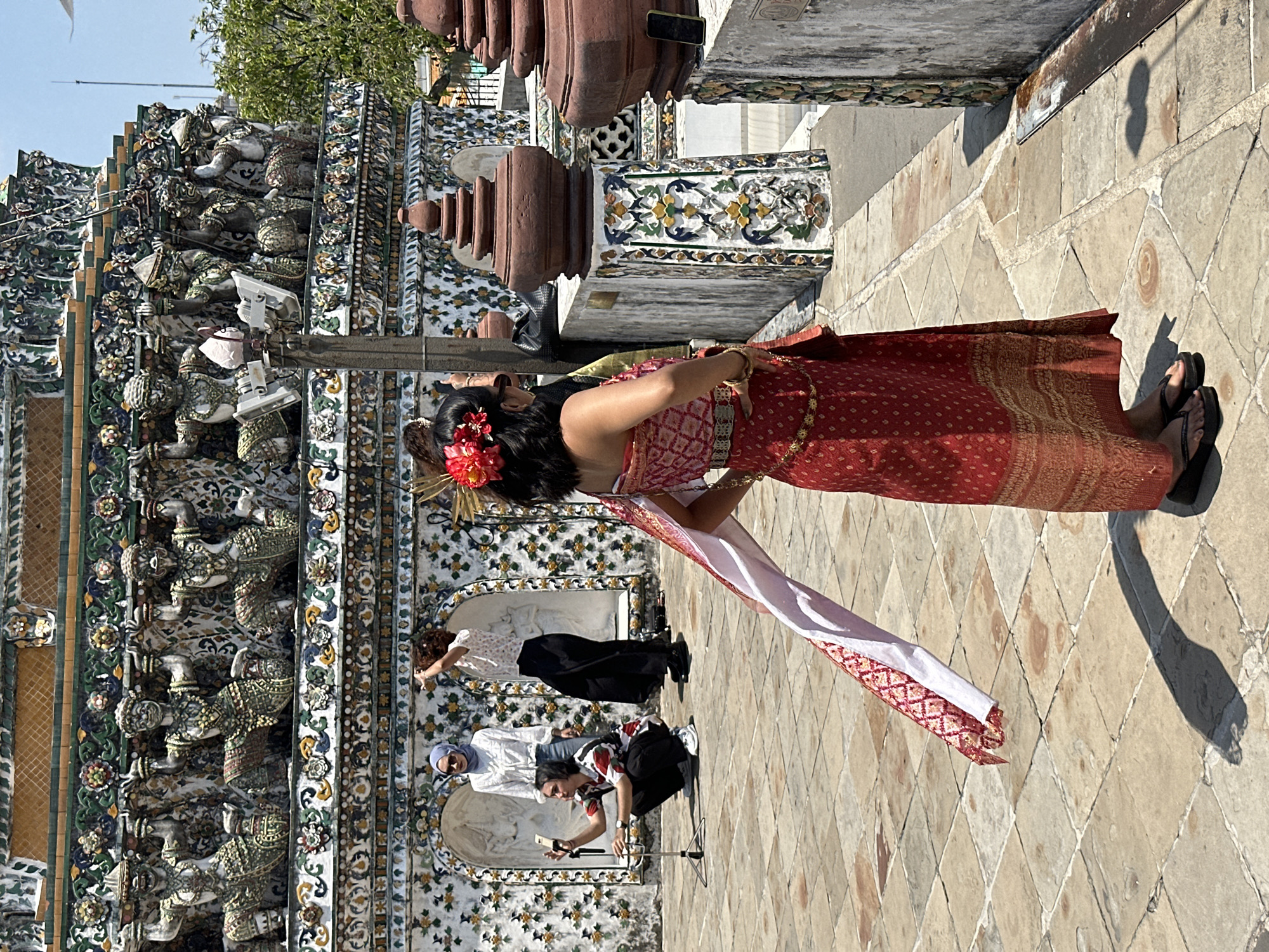 A woman dressed in a traditional red and gold Thai dress stands in front of Wat Arun, a Buddhist temple in Bangkok, Thailand. The temple, adorned with intricate mosaic tiles, rises in the background. A photographer kneels, taking a picture of the woman with a smartphone attached to a tripod. Another woman stands nearby. The ground is paved with diamond-patterned stones.
