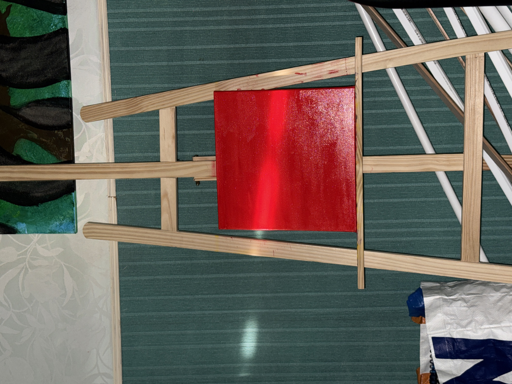 The image shows a red square painting resting on a wooden easel. The easel is holding white cylindrical objects. A painting with dark and green brush strokes is partially visible on the left side of the frame. The background consists of a vertically striped green surface and a patterned white surface. A portion of a blue and white bag is visible in the lower right corner. The location is unknown, and there is no indication of Domont, France.