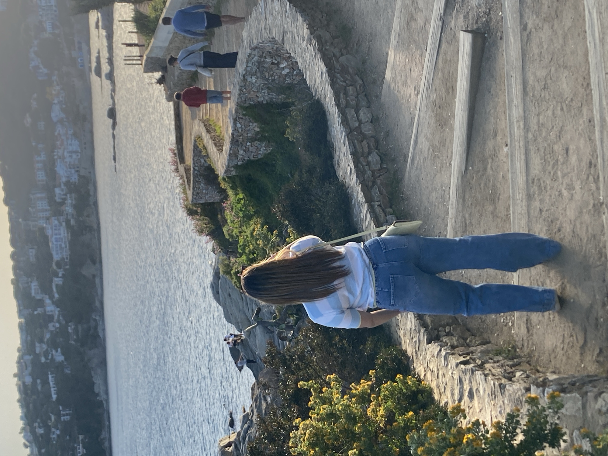 The image shows a scene in Palafrugell, Spain, featuring a coastline view. A woman with long brown hair, wearing a striped shirt and blue jeans, stands on a stone pathway or wall. In the background, a townscape with white buildings is visible across a body of water. Several people walk along a stone staircase further up the path. Vegetation, including green plants and yellow flowers, is prominent along the cliffside. The scene is well-lit, indicating daytime.