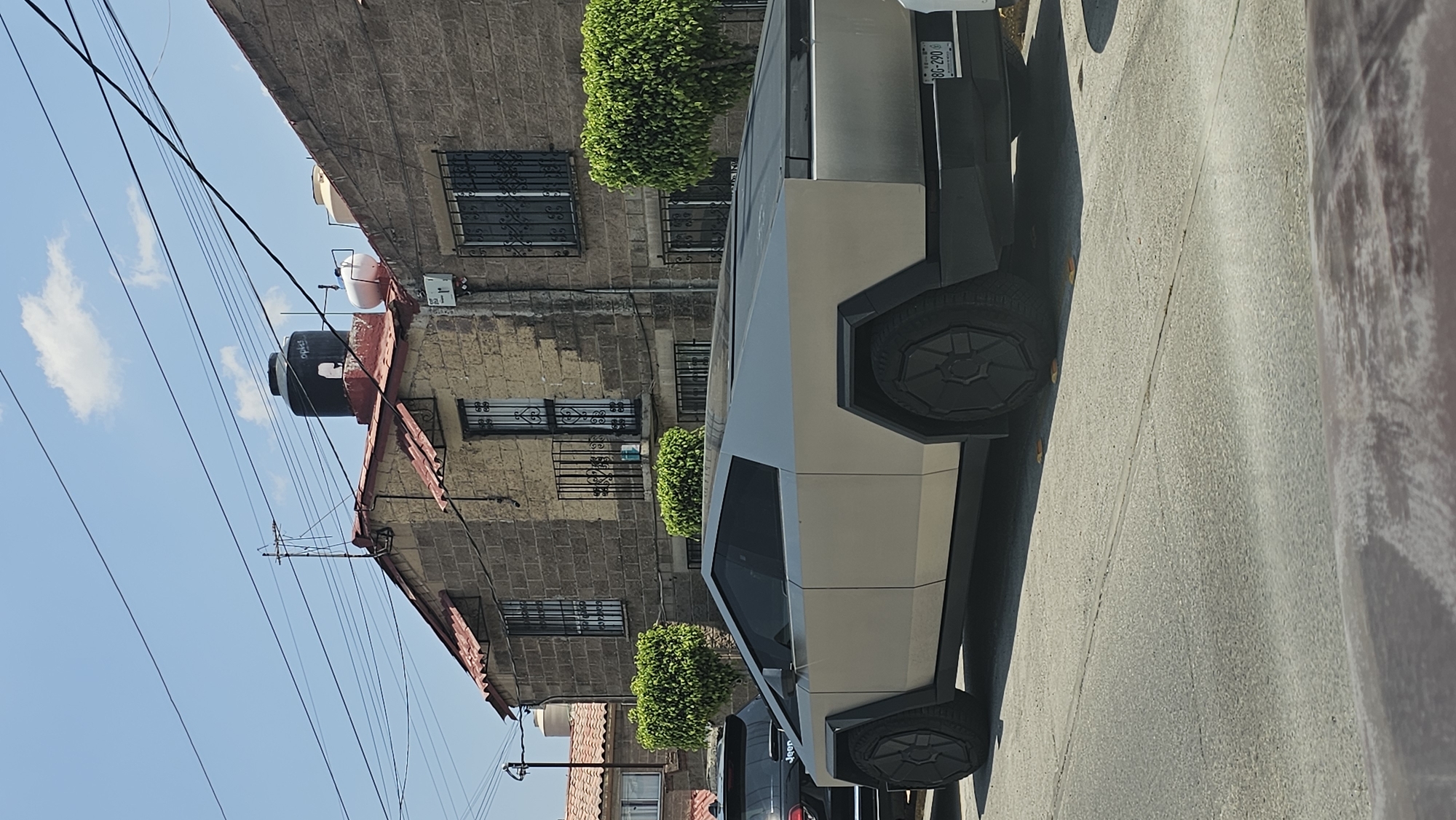 The image shows a Tesla Cybertruck parked on a street in San Bernardino Tlaxcalancingo, Mexico. The truck is silver and has angular lines. It is parked next to a building with stone walls and barred windows. There are bushes in front of the building. The sky is blue with a few clouds. Power lines are visible above the building. The license plate on the truck is visible.