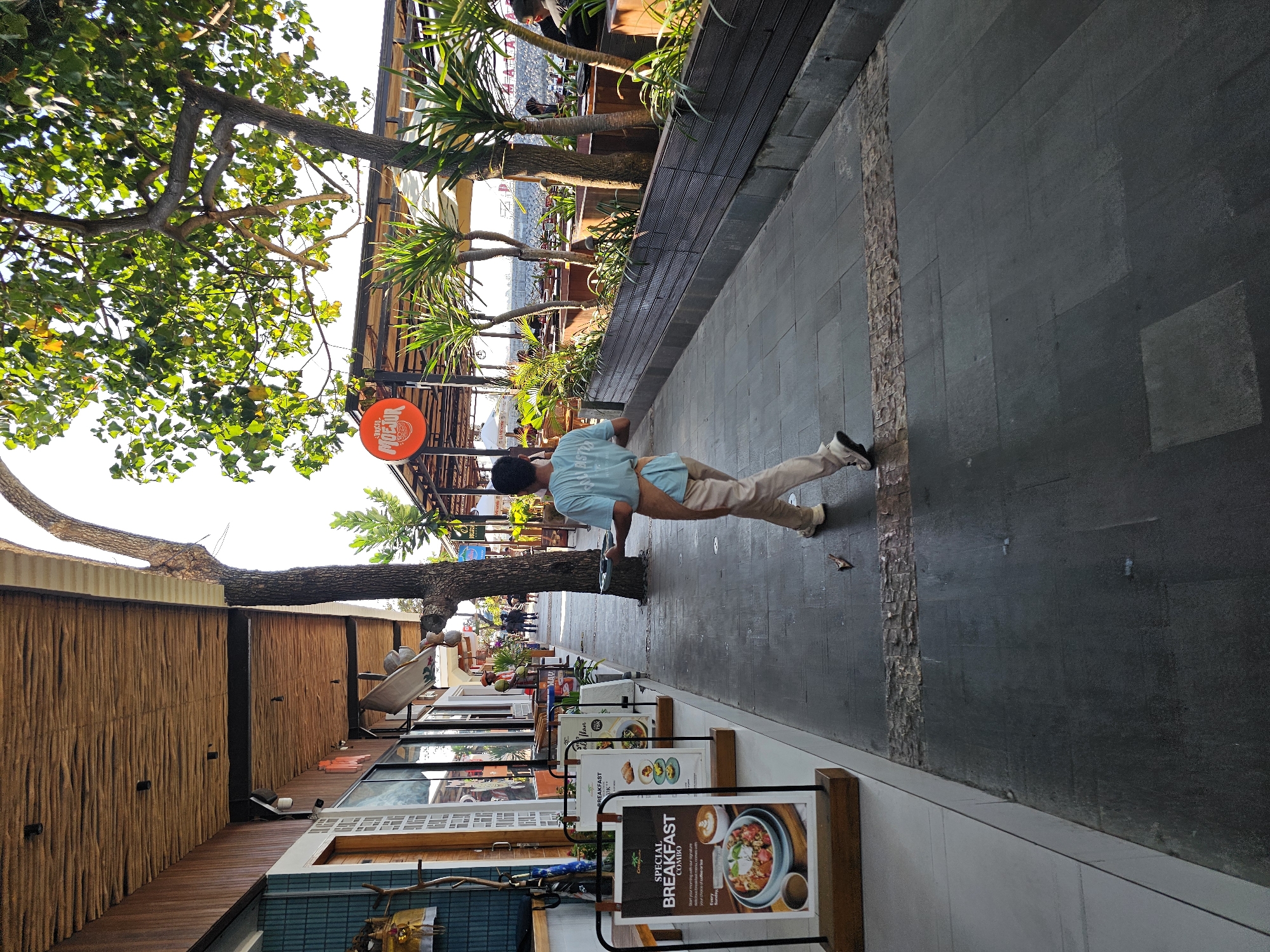 A person wearing a light blue t-shirt and beige pants, with an apron tied around their waist, walks away from the camera holding a silver tray. The person is on a paved walkway next to a row of shops with outdoor seating and foliage. On the left, a menu board displays "SPECIAL BREAKFAST COMBO" with an image of food. The walkway is flanked by a low, dark-colored planter box filled with greenery on the right. In the background, palm trees and other trees are visible, along with a glimpse of the ocean and a beach. The scene appears to be a commercial area in Denpasar, Indonesia, possibly along a beachfront promenade.