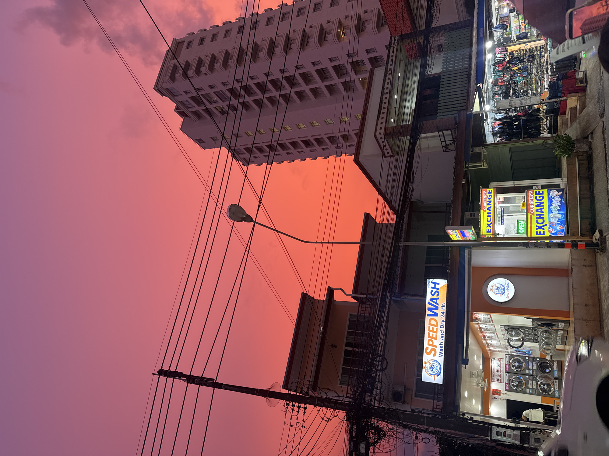 The image depicts a street scene at dusk in Karon, Thailand, under a vibrant pink and orange sky. A tall, white residential building with balconies dominates the right side of the frame, contrasting with a cluster of lower buildings housing businesses on the left. These businesses include a laundromat called "Speed Wash," which is open and illuminated, showcasing washing machines and dryers, and a currency exchange booth. A street lamp with a visible light fixture is positioned between the buildings. A dense network of electrical wires crisscrosses the sky, obscuring parts of the buildings and the sky. The foreground shows the front of a car, partially visible, and the sidewalk. The overall impression is one of a bustling urban environment transitioning into evening, with the dramatic sunset providing a striking backdrop.