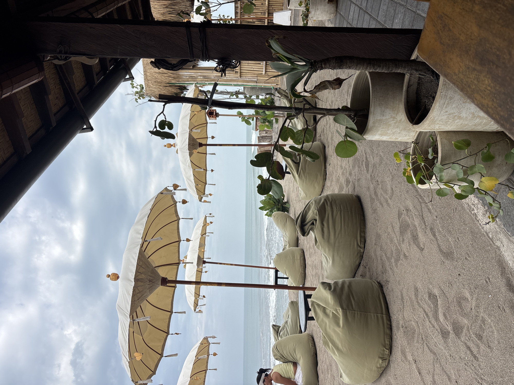 A beachside seating area in North Kuta, Indonesia, features several large, cream-colored umbrellas casting shade over a collection of olive-green beanbag chairs on a sandy beach. The ocean is visible in the background with moderate waves under a cloudy sky. To the right, a covered seating area with a thatched roof and wooden beams is partially visible, with potted plants in light-colored planters placed in the foreground. A person wearing a yellow top and a white visor is seated on one of the beanbag chairs to the left.