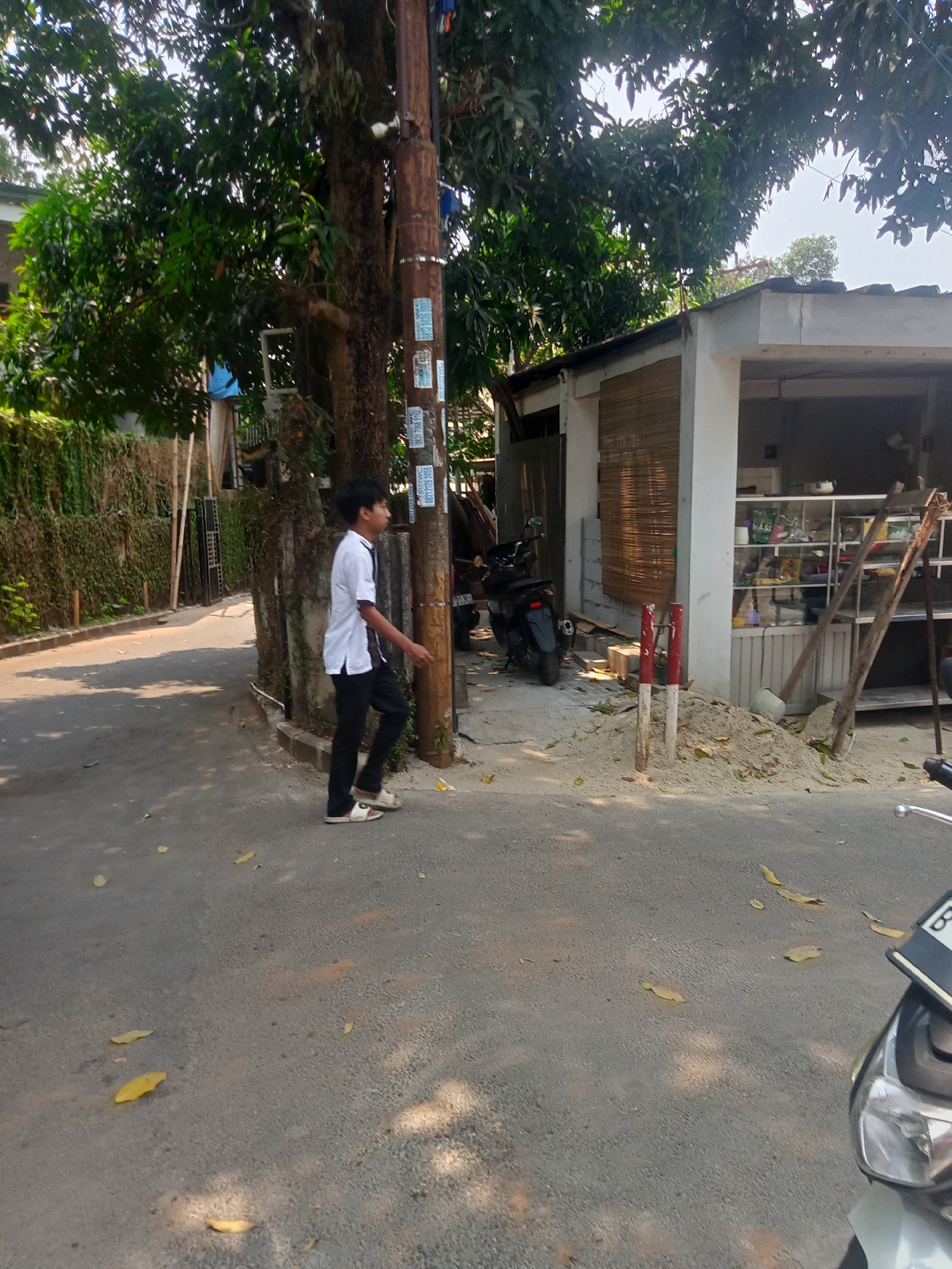 A young male wearing a white t-shirt and black pants walks along a paved street in a residential area of South Tangerang, Indonesia. He is standing to the left of a wooden utility pole, and a green vine-covered wall runs along the left side of the street. Behind him, a small building with a bamboo-covered window and open shelves displays various items. In front of the building, a pile of sand is present, with two red and white striped posts sticking out of it. A black motorcycle is parked to the left of the building, and the front of a white motorcycle is visible in the lower right corner of the image. The ground is littered with fallen leaves.