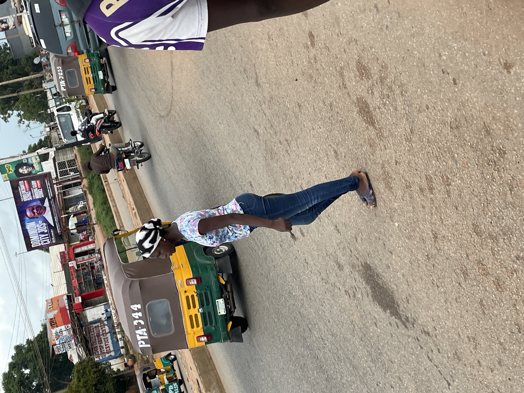 The image shows a street scene in Jos, Nigeria, with multiple vehicles and people. In the foreground, a young woman with a black and white headscarf walks across the street. She is wearing a floral-patterned t-shirt and blue jeans. To her left, a green and yellow auto-rickshaw, identified by the license plate "PTA-344", is parked or moving slowly. Further down the street, another auto-rickshaw with license plate "PTA-3255" is visible, along with a man on a motorcycle. A white van and a green SUV are also present. In the background, buildings with various signs are visible, including a large billboard with an image of a man and the text "DOMINION CITY JOS". The sky is overcast. The road surface appears to be asphalt, with some oil stains and debris.