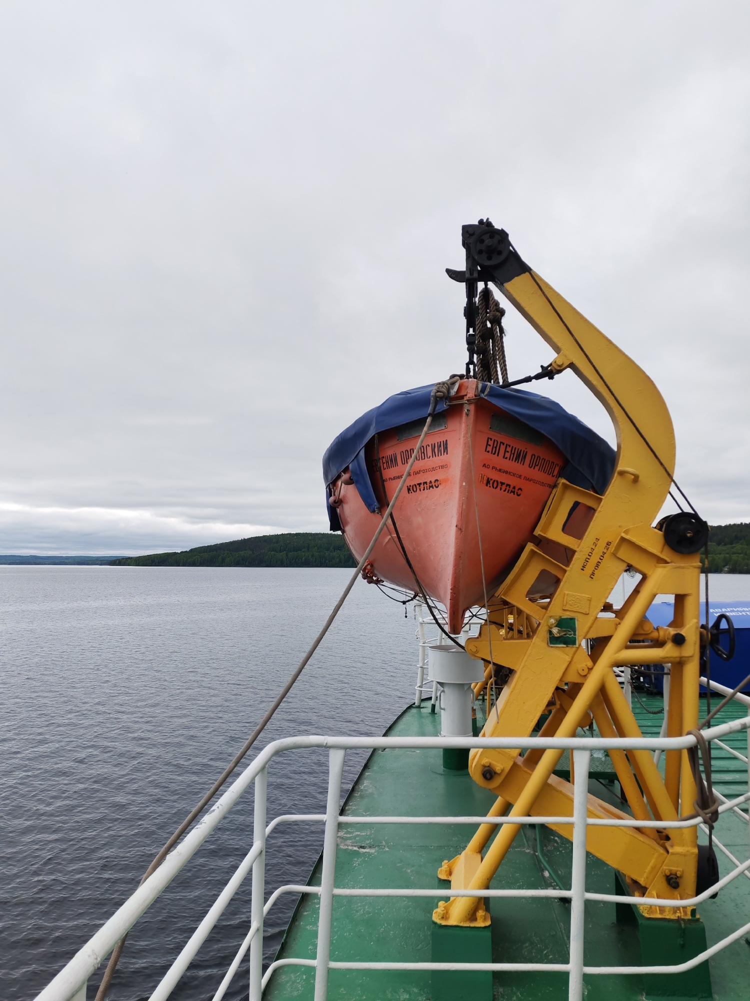 An outdoor wide shot from the deck of a vessel captures a tranquil waterway scene under an overcast sky. In the immediate foreground, a bright orange lifeboat, partially covered with a blue tarp, hangs suspended from a sturdy, yellow metal davit. The lifeboat displays Cyrillic text: "ЕВГЕНИЙ ОРЛОВСКИЙ" (Yevgeny Orlovsky), "АО РЫБИНСКОЕ ПАРОХОДСТВО" (Rybinsk Shipping Company), and "КОТЛАС" (Kotlas), indicating its name, operator, and home port. The yellow davit also has markings "ИСП.04.24" (Inspection/Expiry April 2024) and "ПРОВ.04.26" (Test/Next Test April 2026), suggesting maintenance records.
The green deck of the vessel, with white railings, is visible in the lower portion of the image. The vessel navigates a wide body of calm, dark water that extends to a distant shoreline blanketed by dense green forest. The sky is uniformly cloudy and grey, creating a soft, diffused light over the scene. No people are visible. The setting is likely a large lake or wide river, consistent with the provided location context of Medvezhyegorsk, Russia. The lifeboat's position suggests it is either ready for immediate deployment or recently returned from use/maintenance.