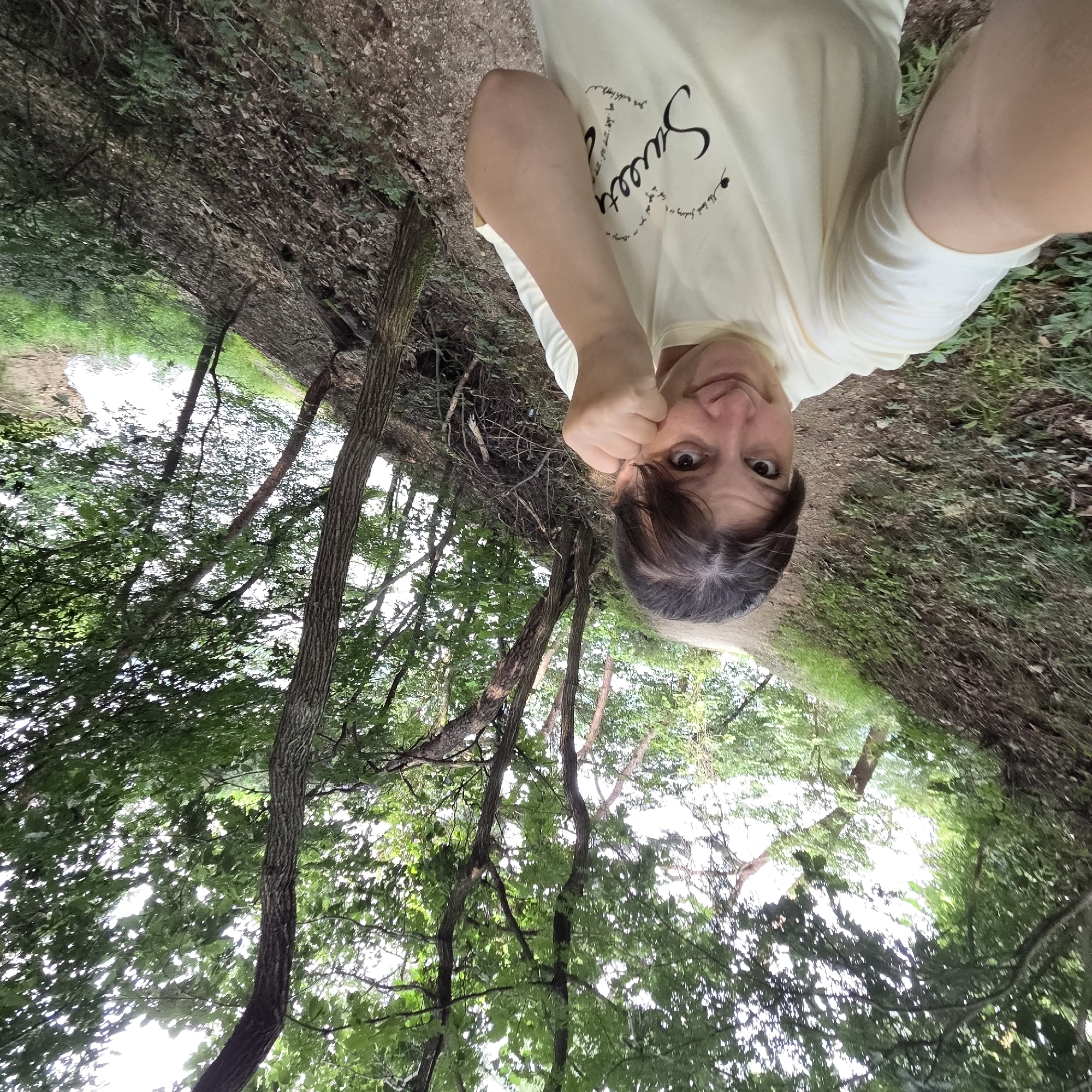 A woman is taking a selfie on a dirt path in a wooded area. She is wearing a light yellow t-shirt with "Sweetest" written in cursive on the front. Her right hand is raised to her face, with her knuckles touching her cheek. She is smiling at the camera. The path is surrounded by trees and greenery, with a steep, leaf-covered incline to her left. The sky is overcast and visible through the tree canopy. The location is Gapcheon-myeon, South Korea.