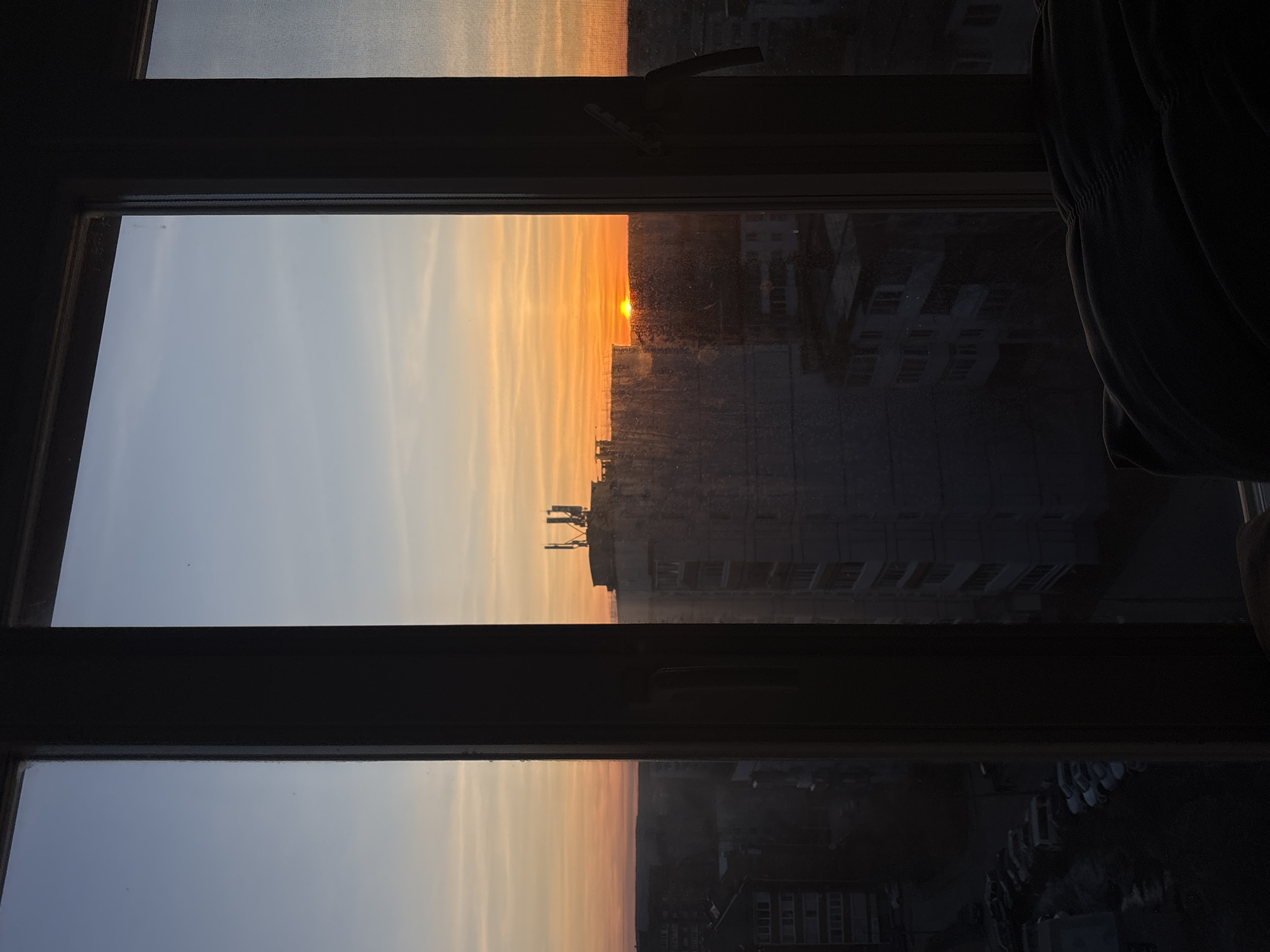 A photograph captures a vibrant sunset or sunrise viewed through a window overlooking an urban landscape in Chaikovsky, Russia. The sun, a bright orange sphere, is low on the horizon, bathing the sky in intense golden and orange hues that fade into a lighter blue and grey. Below the colorful sky, the silhouettes of numerous buildings are visible, their forms indistinct against the encroaching darkness. One prominent building stands out with antenna-like structures on its roof, positioned to the right of the sun.
The dark, thick frame of the window divides the scene, with the main view of the sun and city visible through a central pane. Top and bottom panes reveal more of the sky, along with subtle reflections or smudges on the glass. A dark, pleated curtain or drape is partially visible on the far right, suggesting an interior perspective. The time of day is clearly either dawn or dusk, marked by the sun's low position and the dramatic sky colors. No discernible text is present in the image.
