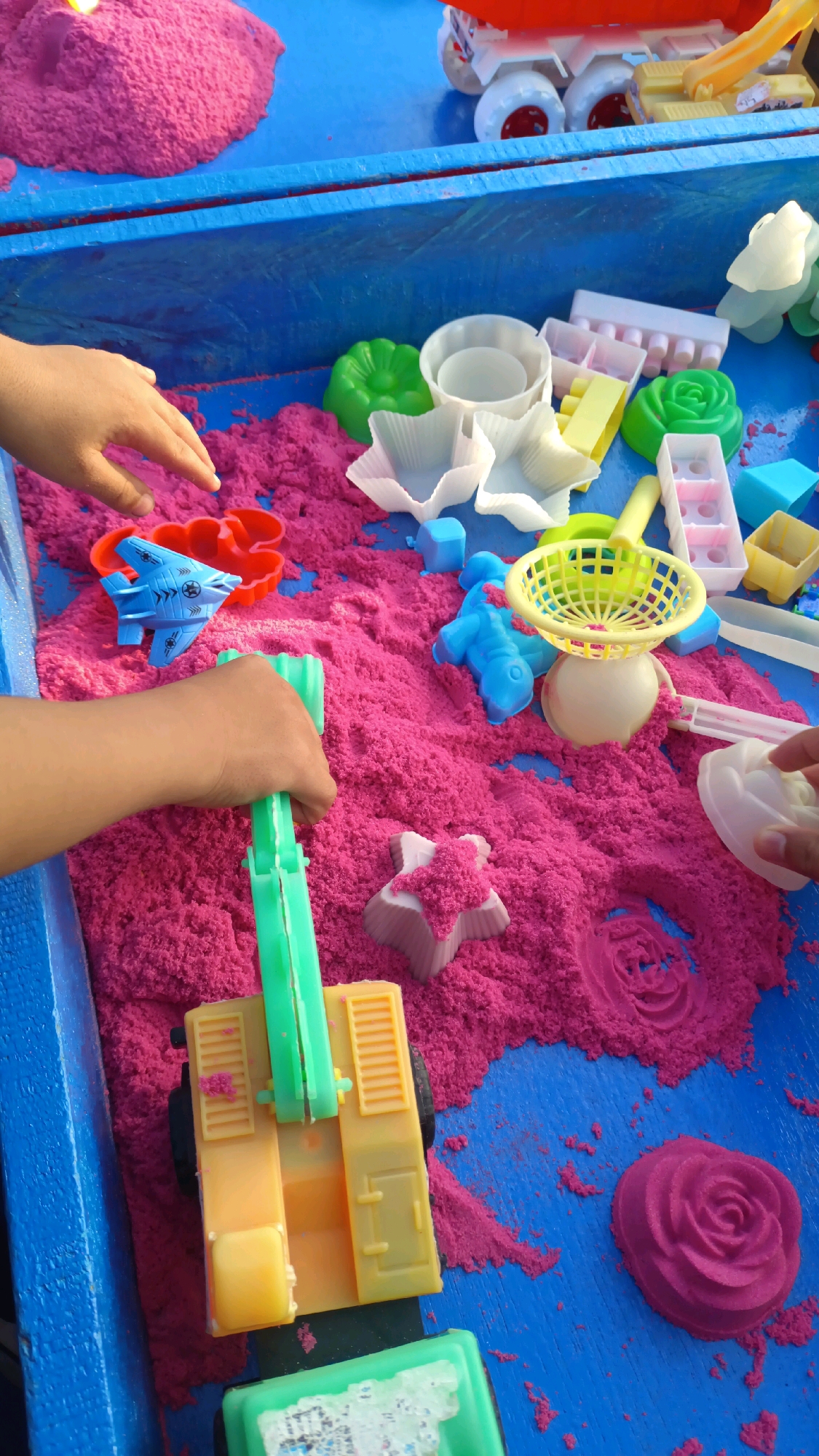 A child's hands are actively engaged with kinetic sand on a blue surface. The kinetic sand is predominantly pink, with some areas of lighter pink or white from molds. Scattered throughout the sand are various plastic toys, including molds in shapes like flowers, stars, and a star-shaped airplane. A yellow toy truck with a green crane arm is positioned centrally, being manipulated by one of the hands. Other toys include blue figures and a yellow basket-like mold. In the background, a red and white toy vehicle with large wheels is visible, partially obscured by the blue surface. The scene appears to be set up for play, likely in an outdoor or recreational area. This activity is taking place in Padang Panjang, Indonesia, where the environment suggests a warm climate suitable for such outdoor play.
