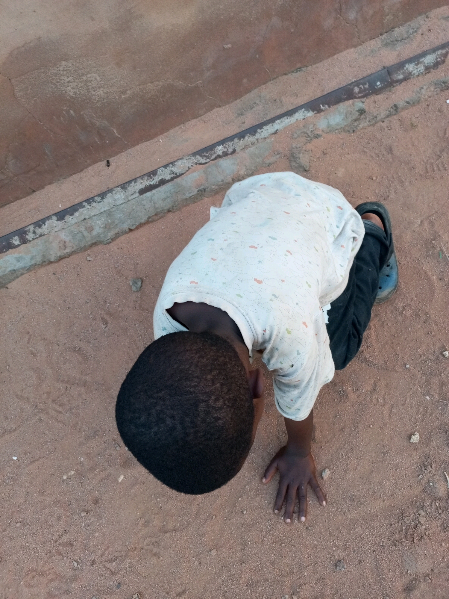 A young Black child is in a push-up position on sandy ground in Tempure, Nigeria. The child is wearing a light-colored shirt with a subtle pattern and dark pants. One hand is on the ground, fingers spread, supporting the child. The ground is reddish-brown sand with some small stones and debris. In the background, there is a textured concrete surface with a thin metal pipe running along its edge. The lighting suggests it is daytime.