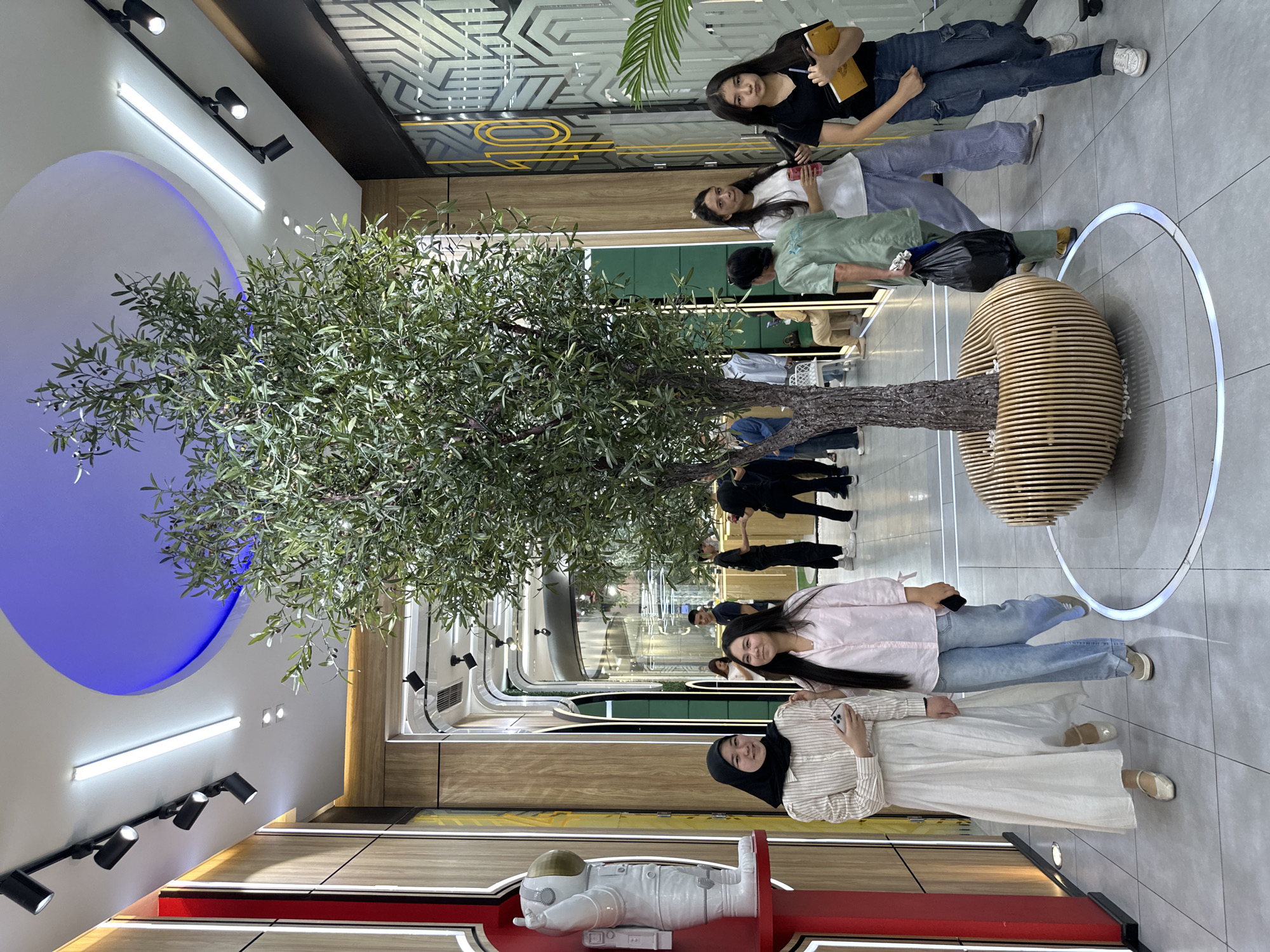 The image shows the interior of an unidentified location in Tashkent, Uzbekistan. A large artificial tree with a wooden base sits in the center of a brightly lit hallway. People, including women wearing hijabs, are present in the hallway, some appearing to be walking or standing. There's an astronaut figurine or statue on a red base in the foreground. The architectural style appears modern with recessed lighting and decorative elements on the walls and ceilings.
