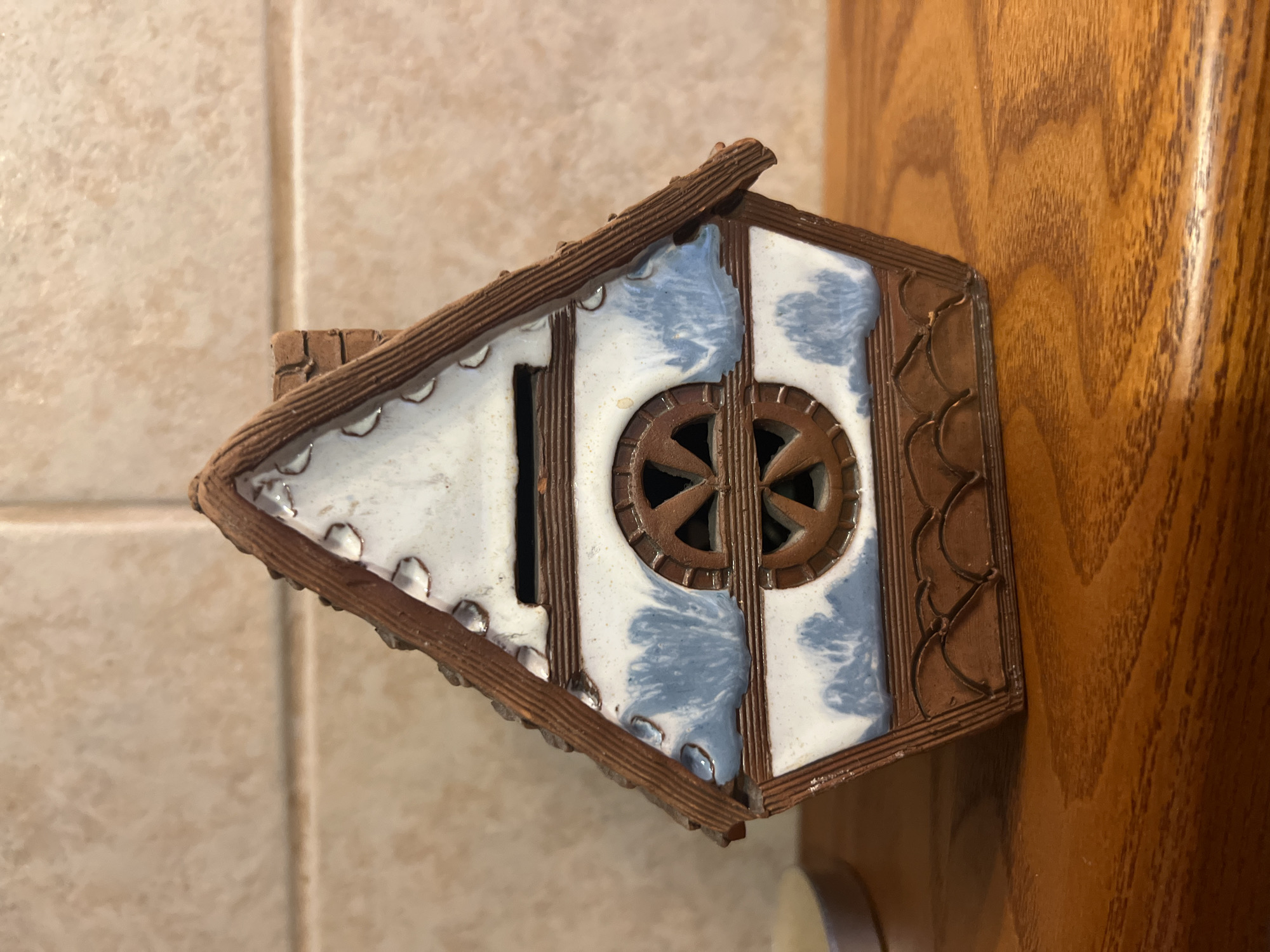 The image shows a close-up, high-angle shot of a small, decorative ceramic house. The house is triangular in shape, with a brown, textured roof that resembles shingles. The front of the house features a white ceramic facade with blue, watercolor-like splatters. There is a small, dark rectangular opening on the left side of the facade, possibly a window or door. In the center of the facade, there is a circular, brown ceramic element designed to look like a wheel with spokes. The edges of the house are outlined with brown ceramic trim. The house is positioned at an angle, leaning against a warm-toned wooden surface, which appears to be furniture or a door. In the background, a beige tiled wall is visible. The lighting suggests it is daytime, with natural light illuminating the scene. The overall impression is one of a charming, handcrafted decorative object. There is no visible text in the image.