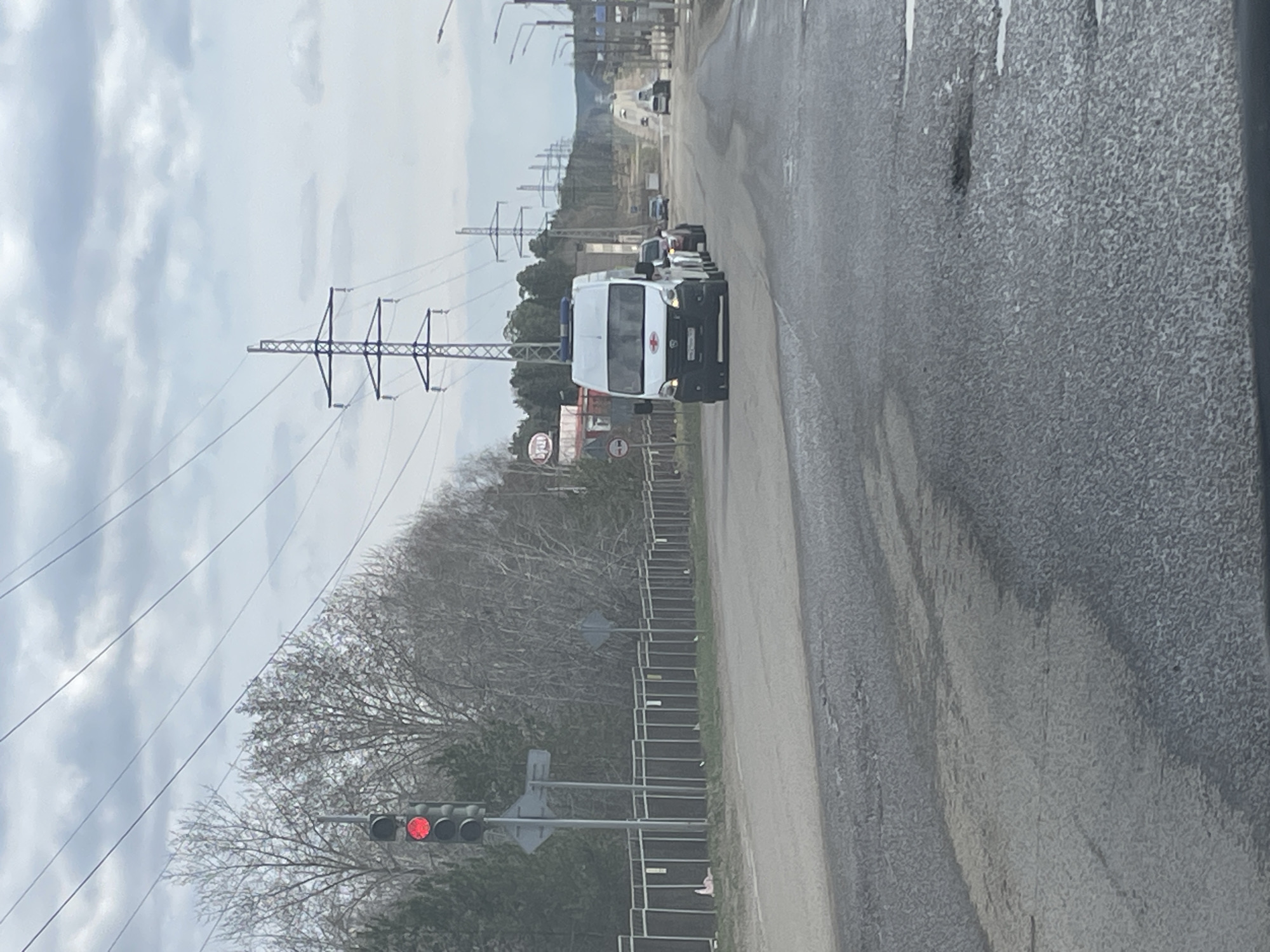The image depicts a white ambulance van with a red cross on its side, traveling on a two-lane road. Power lines stretch across the sky, and trees line the side of the road. A traffic light displays a red light. The scene is located in the city of Chaikovsky, Russia. Other vehicles are visible further down the road. A sign is also visible. A guardrail runs along the edge of the road, with a wooded area and a sky overcast with clouds in the background.