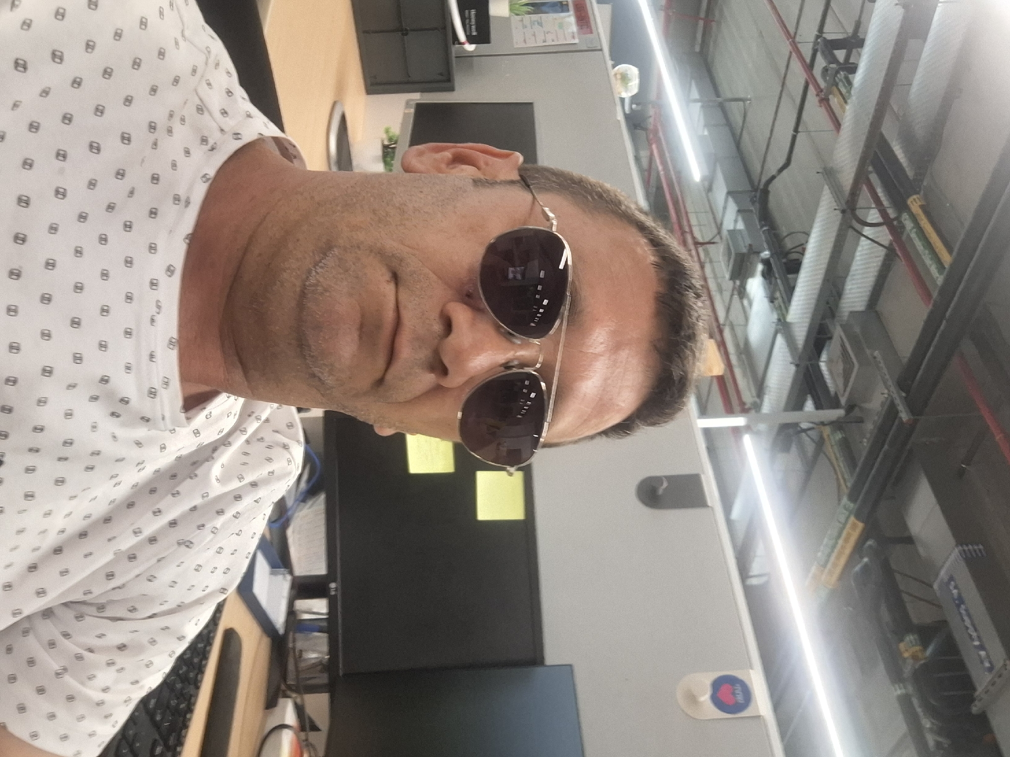 A man wearing sunglasses and a white t-shirt with a repeating pattern of small black squares with a white line through the middle is shown in a close-up selfie. He appears to be indoors, in an office environment. In the background, there are computer monitors, office furniture, and ceiling infrastructure with pipes and lighting. The image is taken from a slightly low angle, looking up at the man's face. The setting does not provide specific visual cues to confirm it is located in Kiryat Gat, Israel.