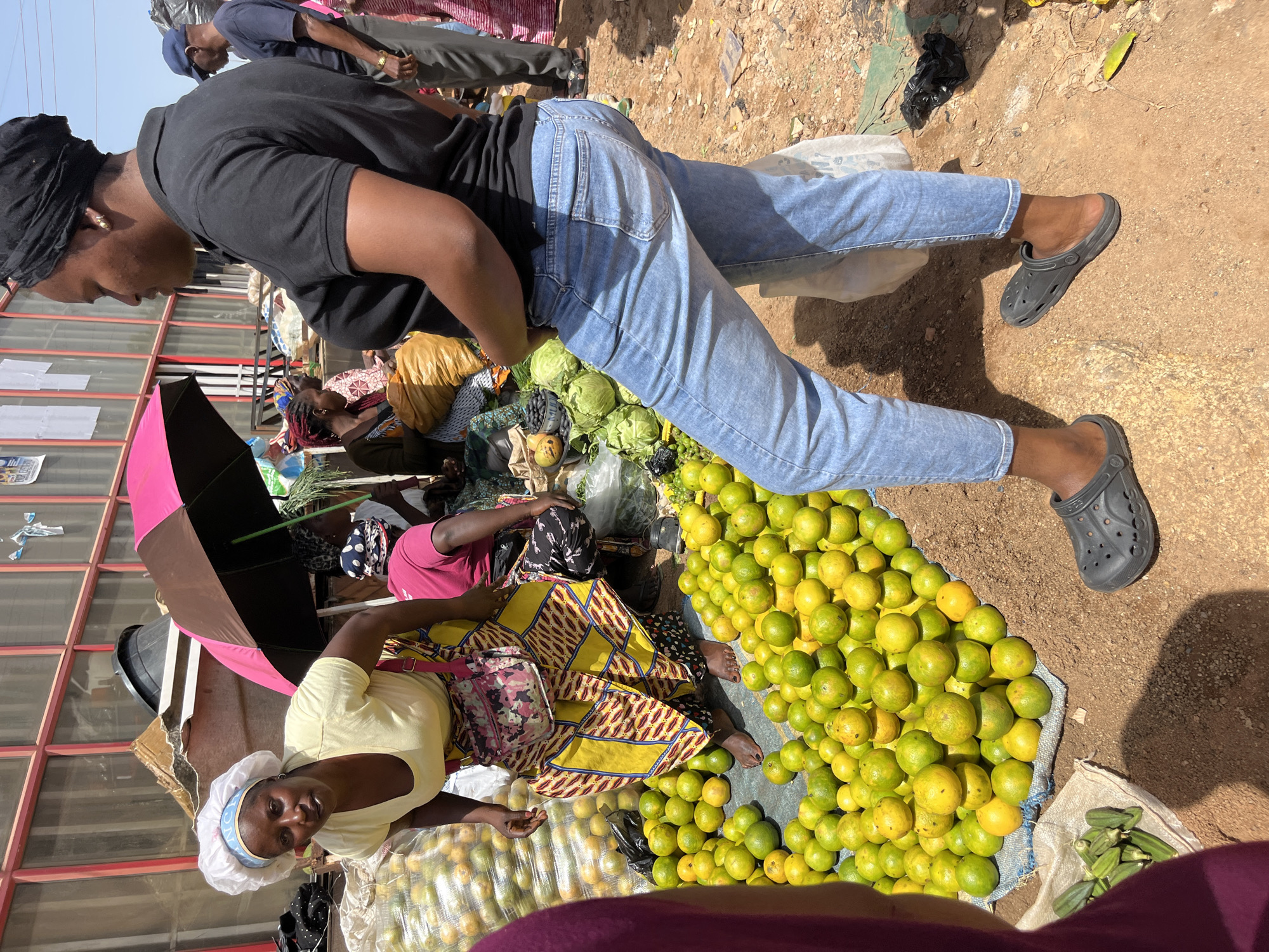 In Gura Top, Nigeria, a bustling outdoor market scene unfolds. Two women, one wearing a yellow shirt and a vibrant patterned skirt, and the other in a black polo shirt and light blue jeans, are at the center of the frame. The woman in yellow is bent over, her head covered by a white cloth, amidst piles of oranges and cabbages. The woman in blue stands tall, looking to her left, with a black headscarf. Their interactions are not visible. The market stalls are laden with produce, including large quantities of oranges, cabbages, and okra, some of which are bagged or piled on the ground. Various people, including a man in a blue cap and another in a pink shirt, are also present, suggesting a busy commercial environment. The ground is dusty, typical of an outdoor market setting. A pink and brown umbrella provides shade for some of the stalls.