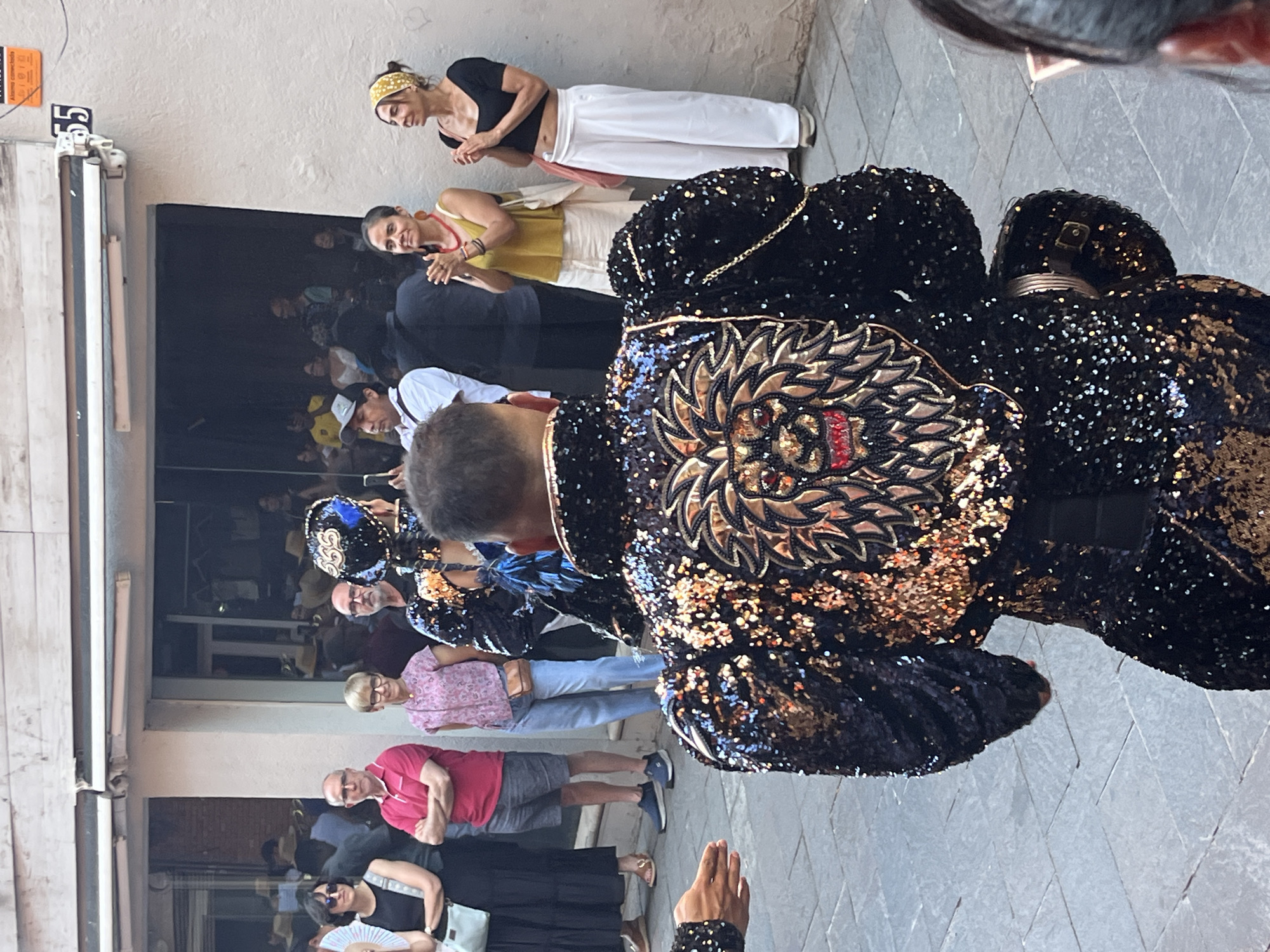 In Sant Cugat del Vallès, Spain, a person in a sequined black jacket with a lion's head design on the back is observed from behind, facing away from the camera. The jacket is adorned with numerous gold and black sequins and features puffed sleeves. Several onlookers stand on a paved street in front of a building with an awning and a glass-fronted establishment. Among the onlookers are a woman in a black dress holding a bag, a man in a red polo shirt and gray shorts, a woman in a pink patterned shirt and blue jeans, a man in a white shirt, and two women in casual attire, one in a yellow tank top and the other in a black crop top and white pants. The scene appears to be outdoors during the daytime, possibly a street event or performance.