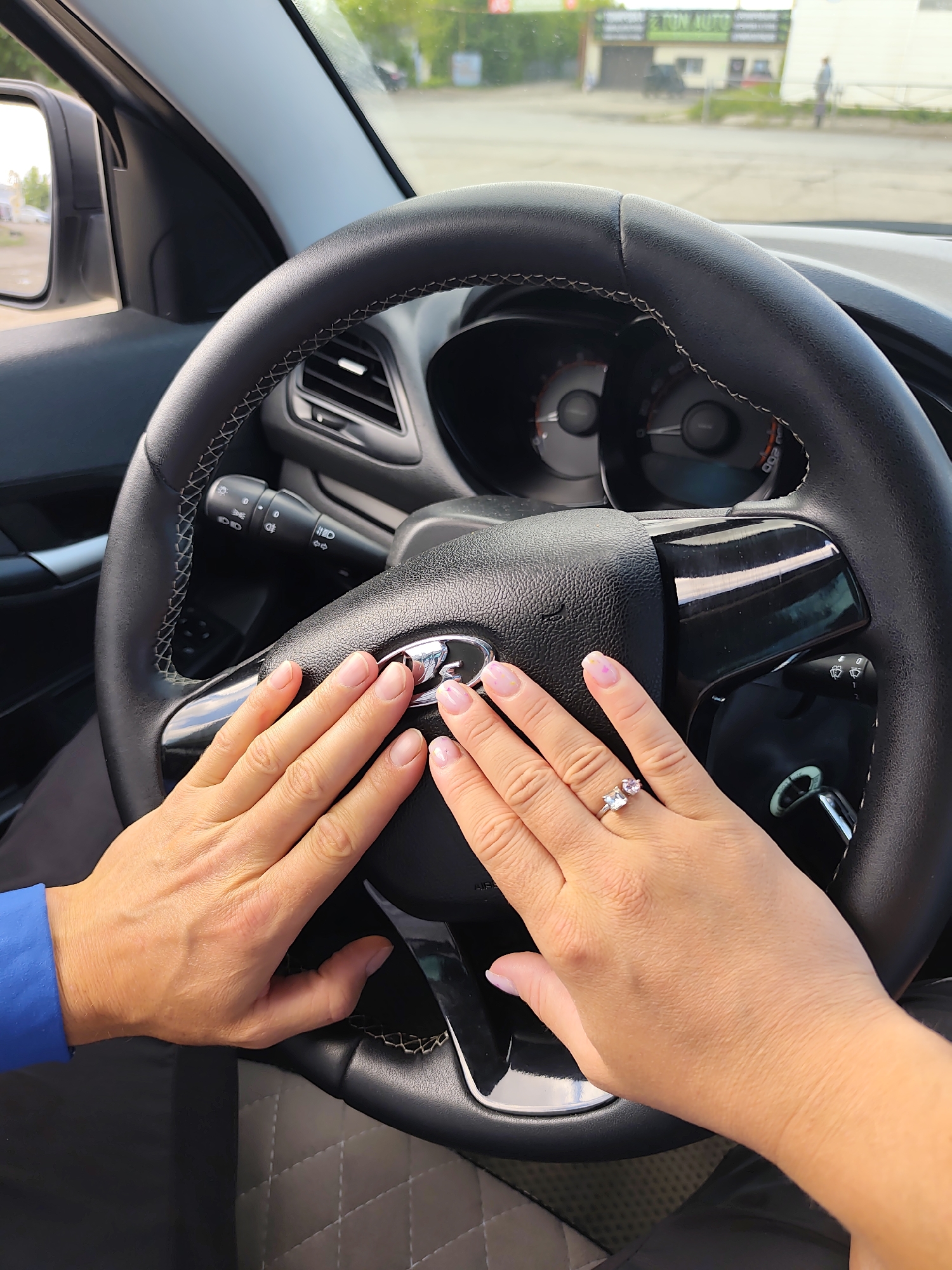 A close-up, interior shot shows a couple's hands resting on the steering wheel of a Lada car. The woman's hand is adorned with a ring. A view through the windshield shows an indistinct street scene. A building with signage "2 TON AUTO" is visible in the background. This points to a location in the city of Chaikovsky, Russia.