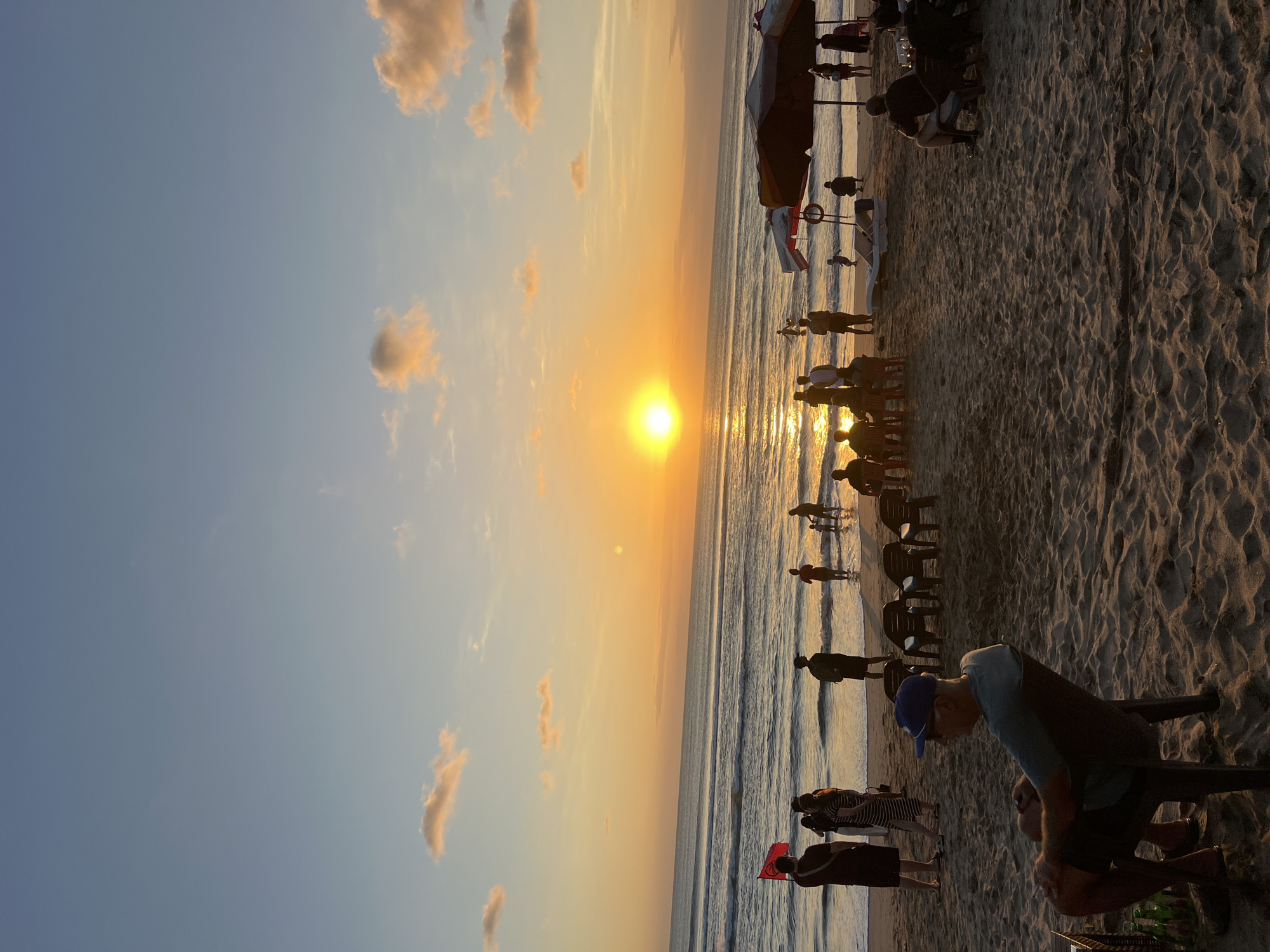 This image depicts a sunset scene at Kuta Beach in Bali, Indonesia. The sun is low on the horizon, casting a warm orange glow across the sky and reflecting on the ocean. Several people are visible on the beach and in the shallow water. Some are standing, while others are seated in chairs. A few individuals are wading in the ocean, and children can be seen playing near the water's edge. Beach umbrellas and chairs are scattered across the sandy shore. The overall atmosphere is one of a relaxed evening gathering on a popular tourist beach.
