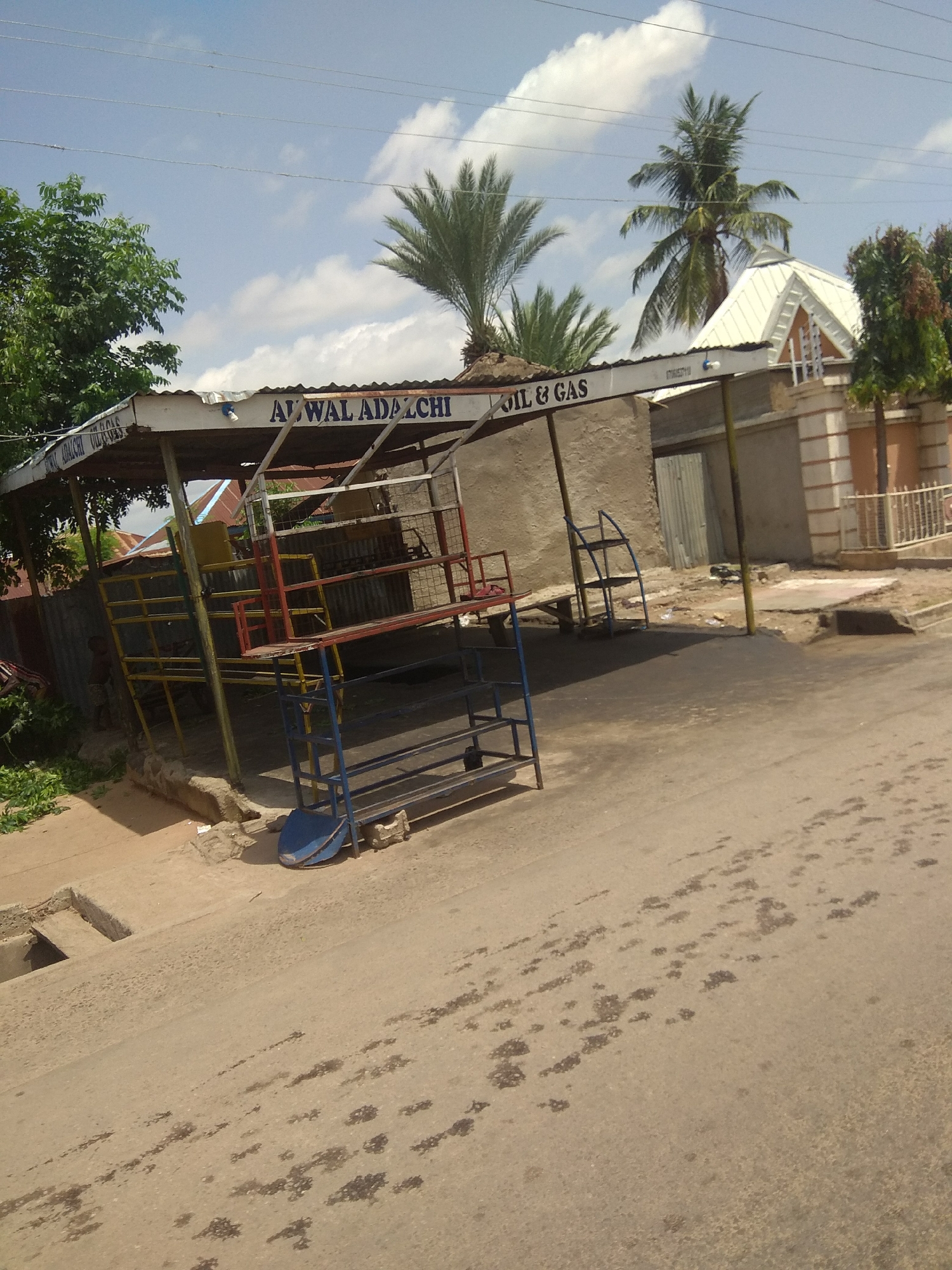 The image depicts an outdoor scene in Jalingo, Nigeria, under a bright, partly cloudy sky. The time of day appears to be morning or afternoon due to the strong sunlight casting shadows. The setting is a roadside area with a small, open-sided structure serving as a business.
The central object is a weathered, open shelter with a corrugated metal roof supported by metal poles. Underneath the shelter, there are several colorful metal racks, possibly for displaying goods or for seating. The structure has signage that reads "AL WAL ADAZCHI OIL & GAS" and "AL WAL ADACHI OIL GAS". To the left of the shelter, a lush green tree provides shade. In the background, a building with a distinctive white, triangular roof and patterned brickwork is visible, along with palm trees and other vegetation. A white metal fence separates the property from the street.
The foreground shows a paved road with dark, irregular splotches, likely oil or water stains, indicating the area's use as an oil and gas vendor. A concrete drainage ditch is visible on the left side of the road. The overall impression is of a humble, functional commercial space in a tropical or sub-tropical environment. There are no people clearly visible in the image. The scene is typical of a small business operating along a road in a Nigerian town.