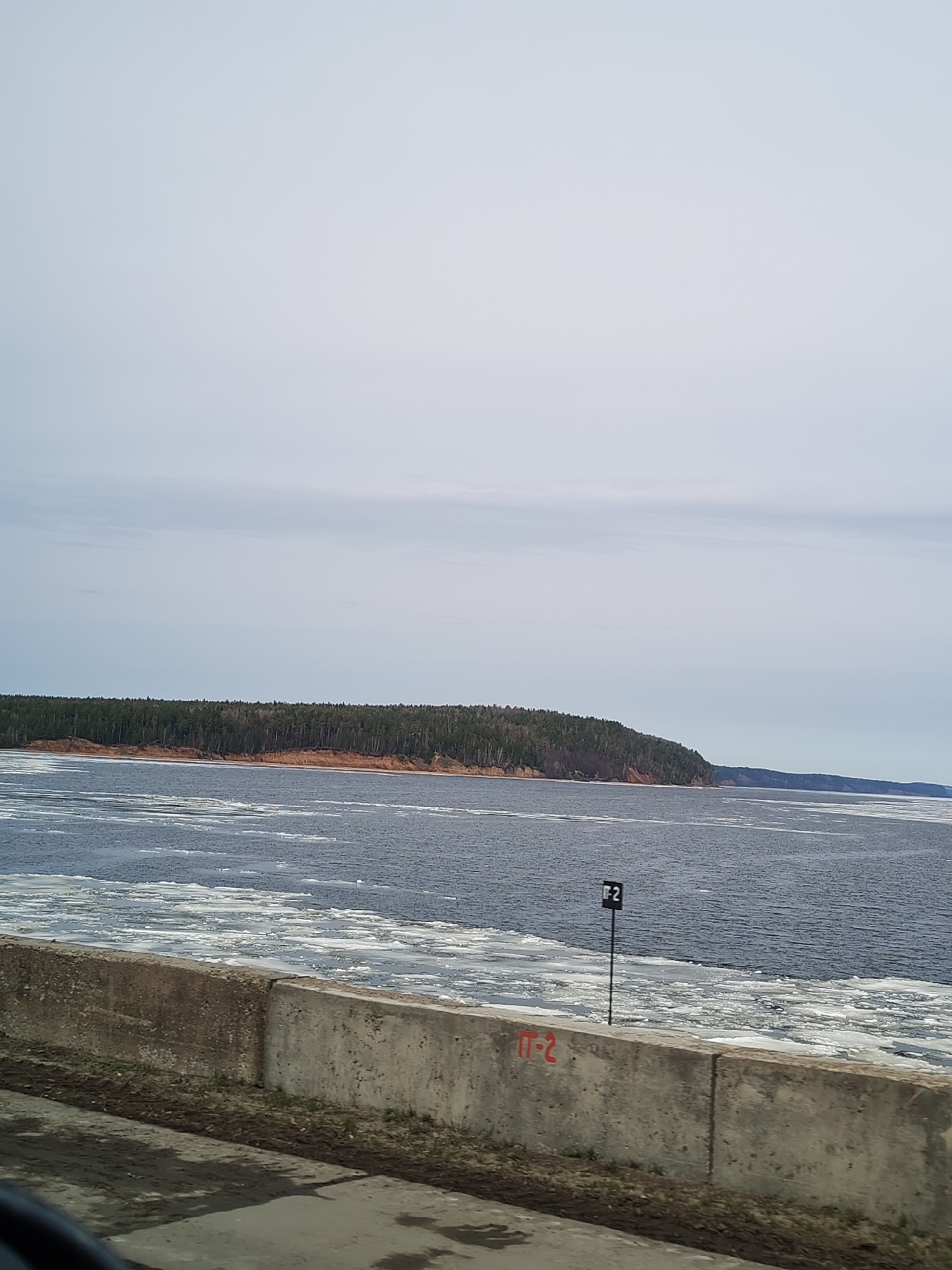 The image shows a view of the Kama River in the city of Chaikovsky, Perm Krai, Russia. The river is partially covered with ice floes. A forested shoreline is visible in the background. A concrete barrier with the inscription "П-2" in red is in the foreground, along with a signpost with the same inscription. The sky is overcast. The overall impression is one of a cold, late-winter or early-spring day.