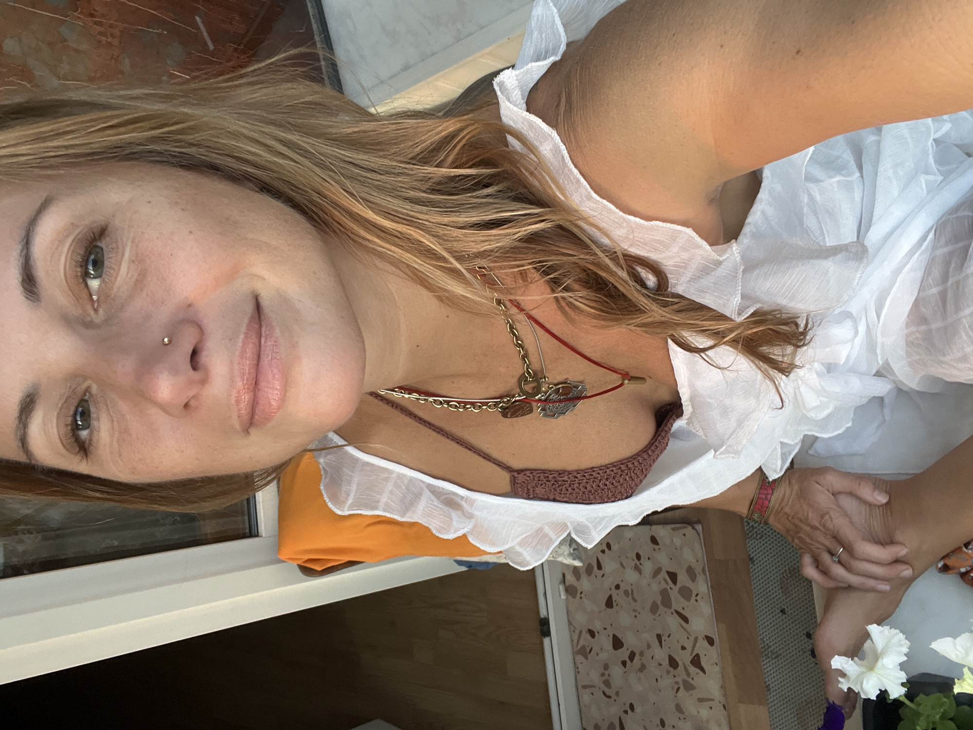 In this close-up selfie, a woman with blonde hair and a nose ring looks towards the camera. She is wearing a white ruffled dress over a brown crocheted bikini top and several necklaces. Her right hand is resting on her left foot, which is adorned with a ring and a red bracelet. She is seated outdoors on a chair with an orange cushion. In the background, a door and a tiled floor are visible. There is no information in the image to determine the location in Samara, Russia.
