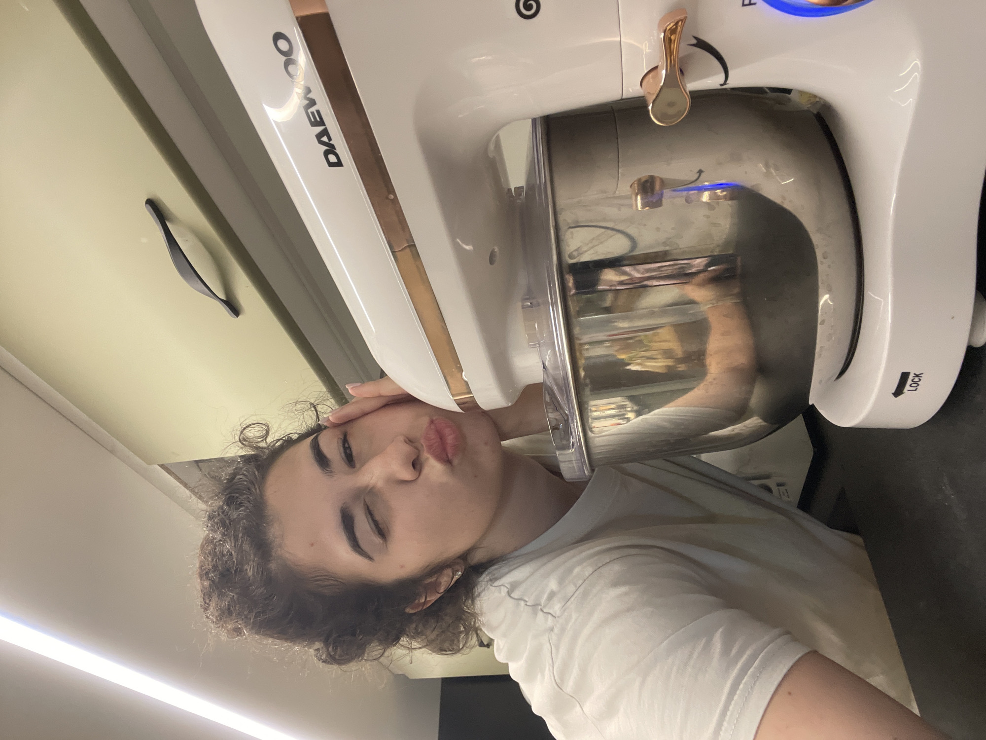 The image displays a playful indoor scene, likely set in a kitchen in Sofia, Bulgaria. A young person, appearing female, with curly dark hair and light skin, is featured prominently on the left side of the frame, puckering their lips in a kissing gesture towards a stand mixer. They are wearing a plain white t-shirt, and one hand rests on the side of the mixer while the other is near their head. Their eyes are partially closed, contributing to the lighthearted expression.
The main object of focus, besides the person, is a white and rose gold/copper DAEWOO stand mixer, positioned on what appears to be a dark countertop or surface. The mixer features a clear plastic splash guard over its metallic mixing bowl. A control knob with a visible blue light and a lever mechanism, also in rose gold/copper, are on the side of the mixer's head. The brand name "DAEWOO" is clearly visible in black text on the white body of the appliance, and the word "LOCK" with an arrow symbol is visible on a white circular mechanism near the base of the mixer.
In the background, a light-colored wall or cabinet is visible, along with a black, curved handle of a drawer or cabinet. Bright overhead lighting is also evident. The overall mood of the image is humorous and affectionate, highlighting a moment of playful interaction with a kitchen appliance. The image is originally captured in portrait orientation but is displayed rotated 90 degrees counter-clockwise.