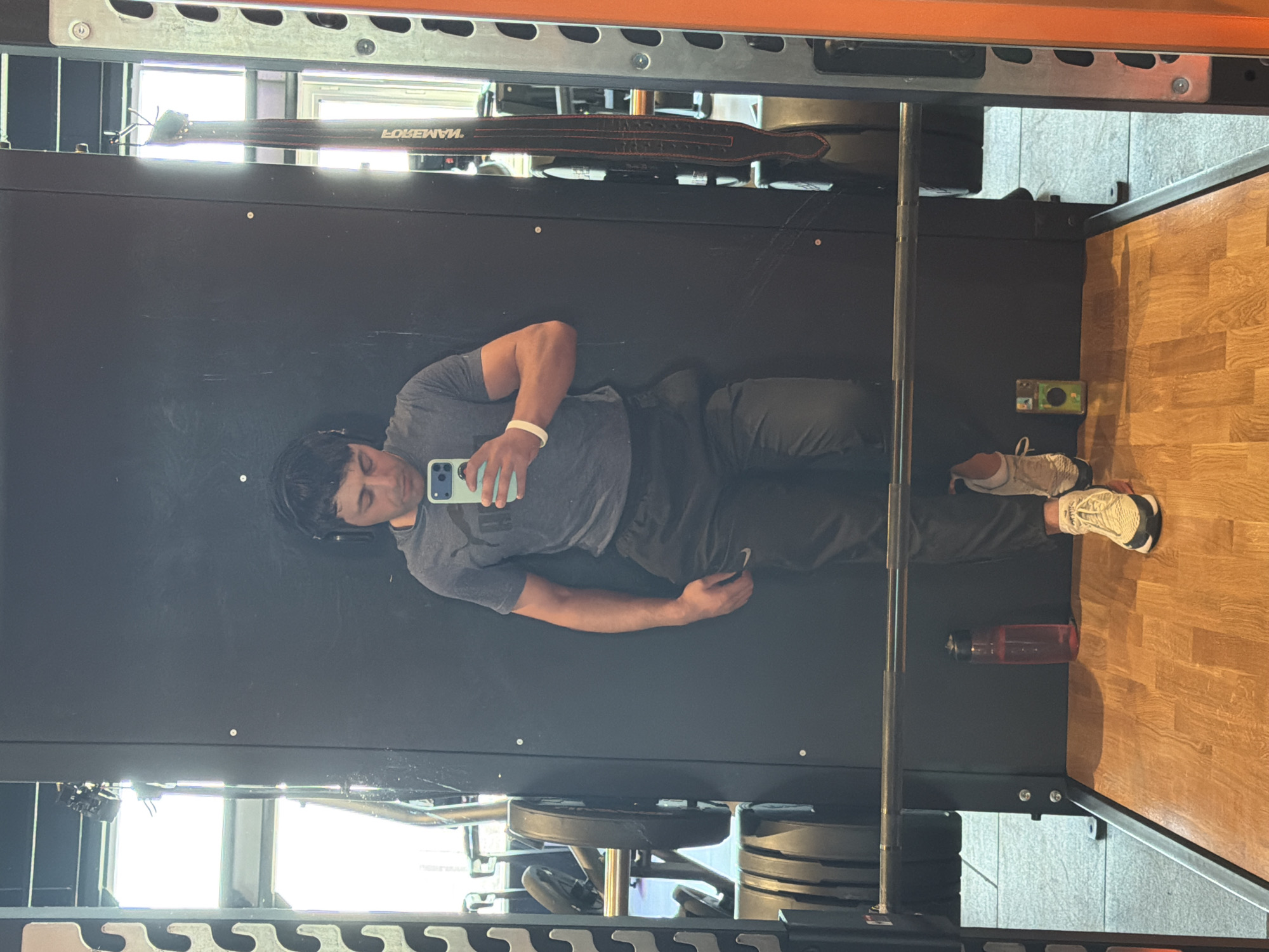 The image is a mirror selfie taken by a man in a gym, depicting his reflection in a large, dark reflective surface, likely part of a power rack or a large mirror. The man appears horizontally as if lying down. He is wearing a gray short-sleeved t-shirt, dark athletic pants, white sneakers, and black headphones. A white wristband is visible on his left wrist, and he holds a light blue/green cased smartphone, seemingly used to capture the image. His expression is neutral.
Above the man's reflection, a black weightlifting belt with red stitching is hanging, showing the reversed text "LOREMAN". Further up, an orange metal beam of the gym equipment is visible. To the right, a wooden-patterned floor extends, with a red water bottle placed near the man's feet. A small green and yellow box-like object is also seen on the dark surface. In the background reflection, a large window is visible, indicating natural light and suggesting it is daytime. Stacked weight plates are partially visible to the left, reflecting in the lower part of the image. The scene conveys a moment during a workout in a gym setting in Krasnoyarsk, Russia.
