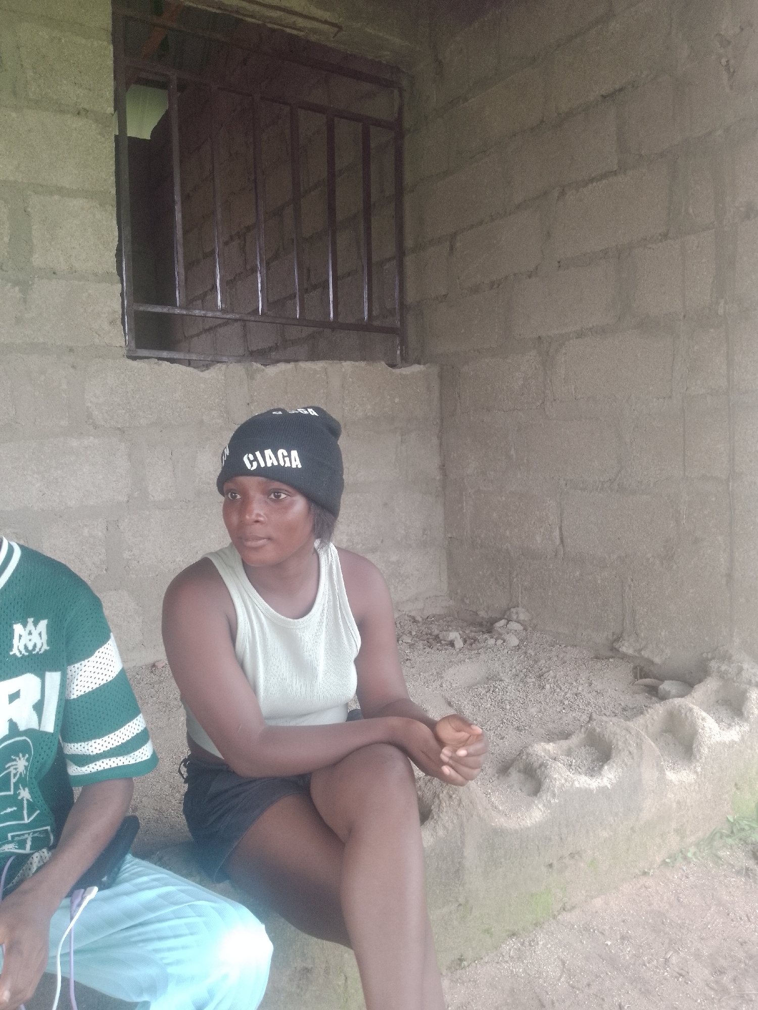 The image depicts an outdoor scene with two individuals in what appears to be Jauro Yinu, Nigeria. On the right, a young woman wearing a black beanie with "CIAGA" written in white, a light-colored tank top, and dark shorts is seated. She is looking to her right, with her hands clasped in her lap. To the left of the woman, a person wearing a green and white striped jersey and light blue pants is visible from the chest down. This individual is holding a black object, possibly a phone, and has white and purple cords extending from it.
The environment features a rough, concrete-block wall forming the background, with a window covered by a metal grate. The ground is covered in sand and small stones. In the foreground, there's a textured concrete structure with indentations, possibly a seating area or a decorative element. The lighting suggests it is daytime.