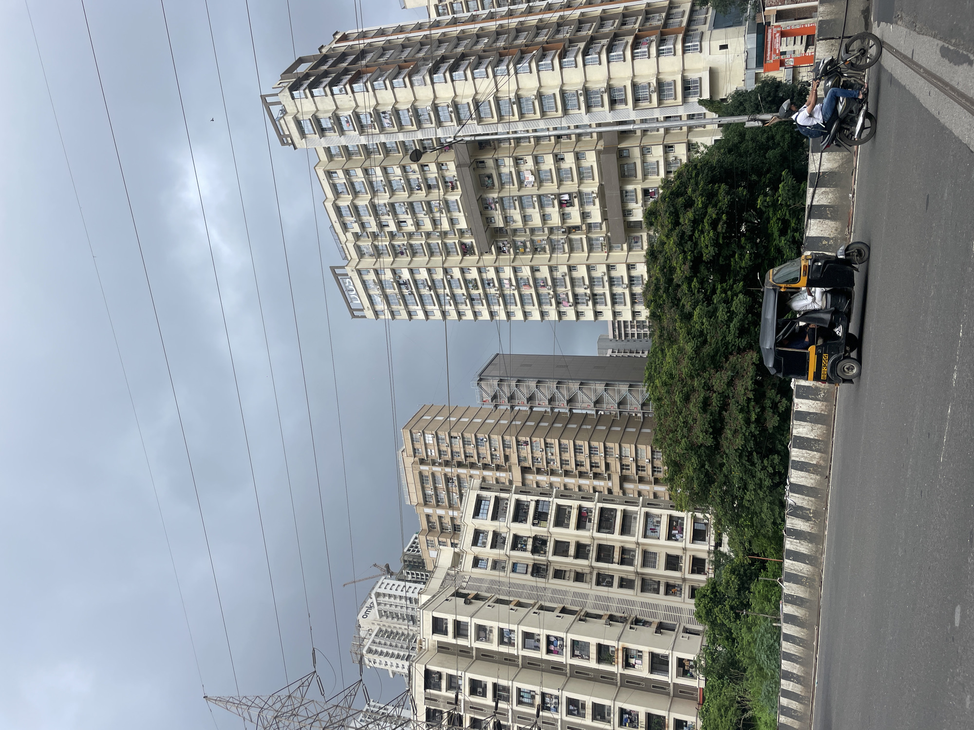 In Mumbai, India, on a cloudy day, a street scene unfolds with towering residential buildings dominating the background. The buildings are modern, with numerous windows and balconies, indicating a densely populated urban area. A prominent structure features the name "DOS" at its top. In the mid-ground, a lush green canopy of trees partially obscures the lower portions of the buildings. The foreground showcases a road with a concrete barrier marked with black and white stripes. Traffic is present, with an auto-rickshaw carrying passengers and a motorcyclist wearing a helmet and backpack. Overhead, power lines stretch across the sky, and a large electricity pylon stands tall to the left, suggesting an active power infrastructure within the city.