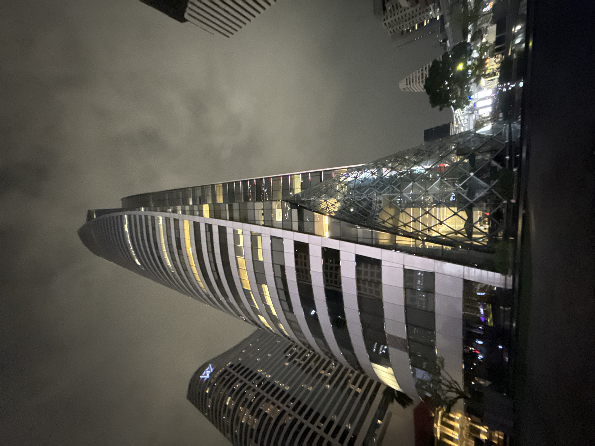 This image captures a nighttime view of the W Bangkok Hotel and the adjacent skyscrapers in the Sathorn district of Bangkok, Thailand. The W Bangkok Hotel, with its distinctive curved facade and illuminated windows, is the central subject. To its left, another skyscraper, likely the adjacent building, is also visible with its own illuminated windows. A prominent feature in the foreground is a uniquely shaped glass structure, part of the W Bangkok complex, reflecting the surrounding lights and sky. In the background, other city buildings, some with visible lights, and trees can be observed. The sky is overcast, contributing to the ambient nighttime lighting. The street in the foreground is dark, with some reflections of lights on its wet surface, suggesting recent rain. No people are clearly visible in the image.