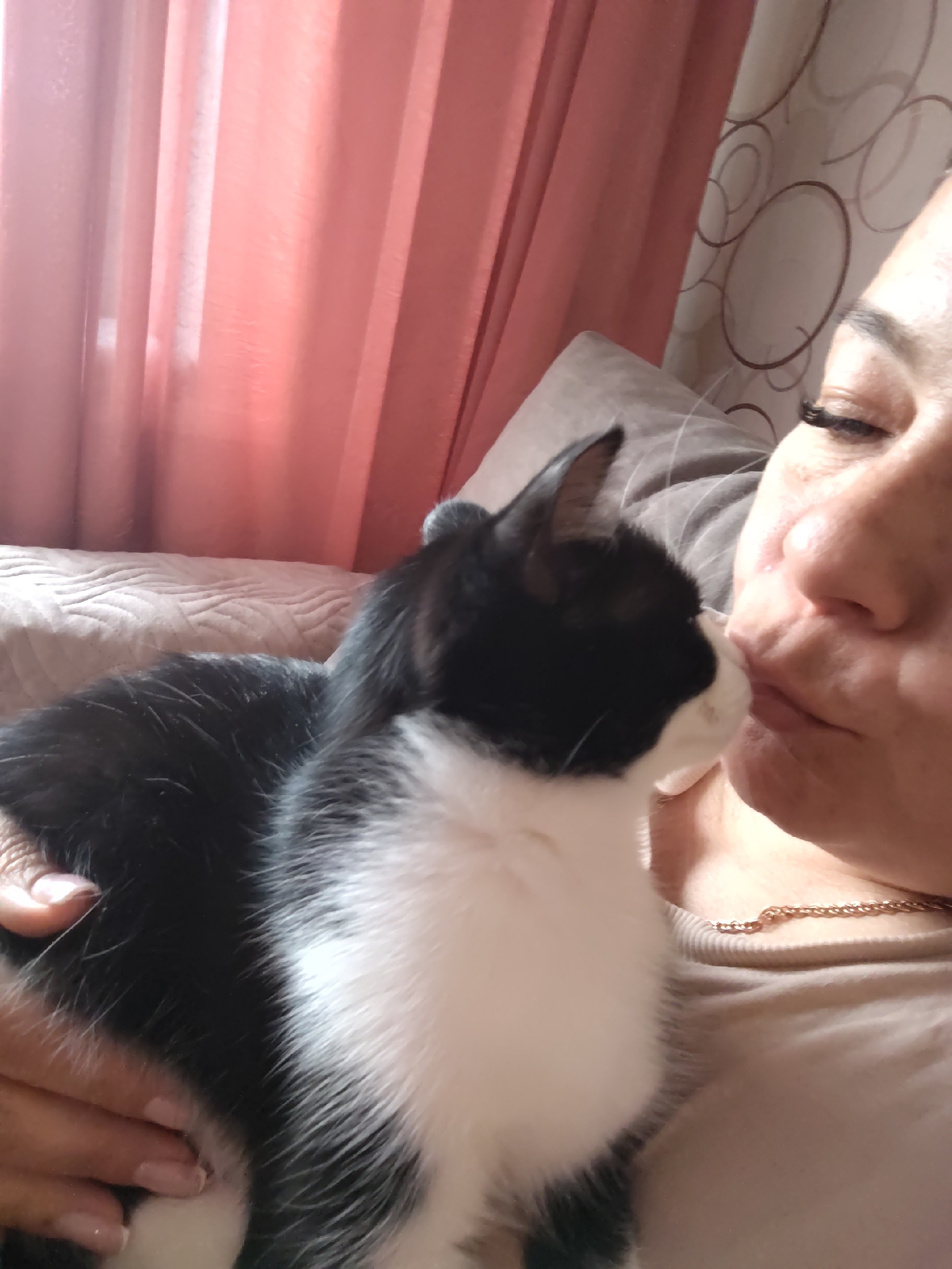 A woman is holding a black and white cat close to her face. The cat's head is near the woman's mouth, and they appear to be kissing or nuzzling. The woman is wearing a beige top and a gold necklace. The background includes a beige pillow, pink curtains, and patterned wallpaper. The location is a room in a house or apartment.
