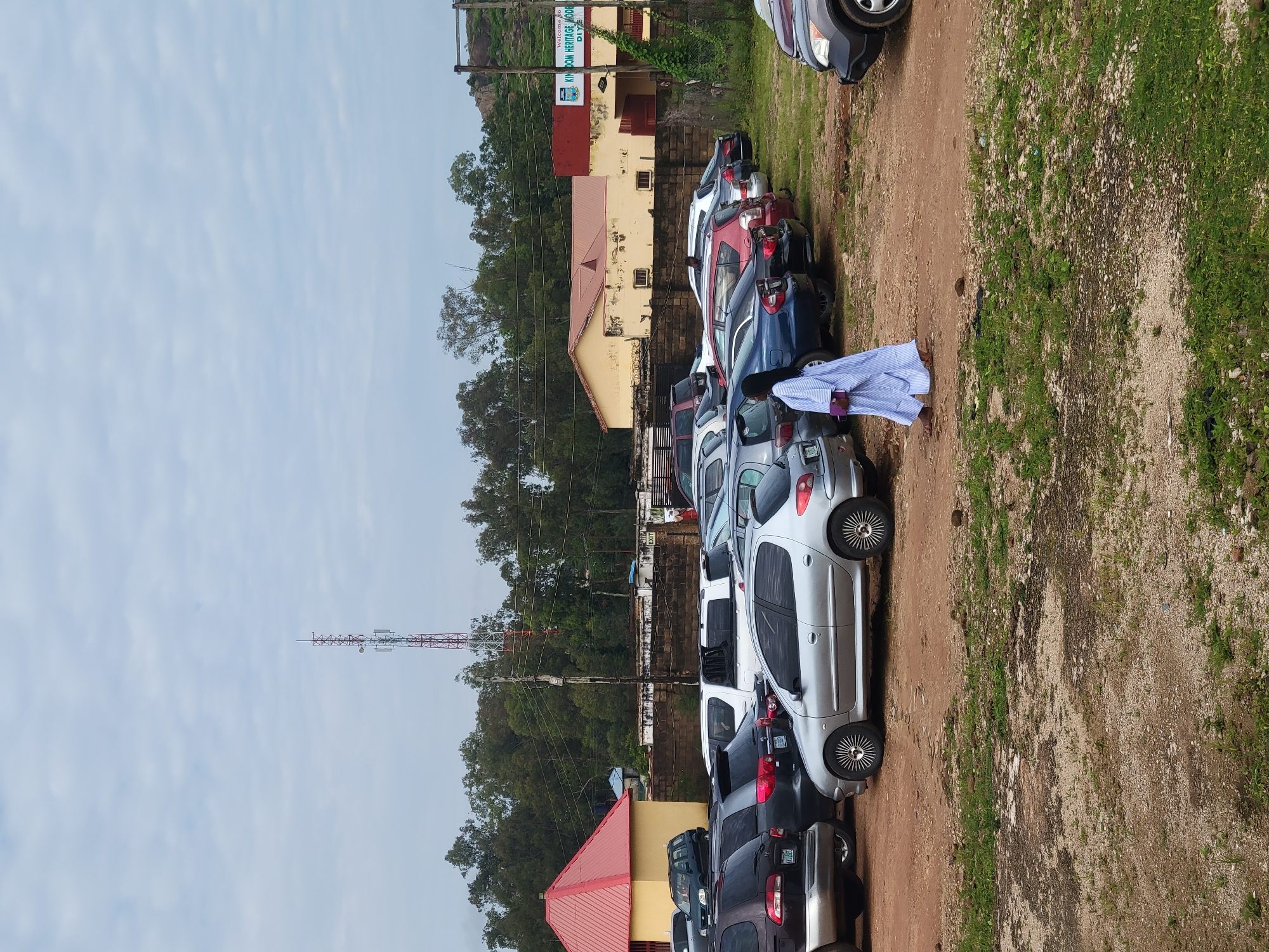 The image depicts a parking lot filled with numerous cars, including silver sedans, dark SUVs, and blue cars. In the mid-ground, a person wearing a light blue and white striped garment is walking across the dirt and gravel parking area. Behind the cars, there is a low brick wall and several buildings, one with a red roof and another with a red and yellow facade. A sign on the latter building reads "Welcome to KINGDOM HERITAGE MODE DIYA". Further in the background, a tall telecommunications tower with red and white sections stands against a cloudy sky. Lush green trees and vegetation are visible throughout the scene, and a rocky hillside is present in the far right background. The overall environment appears to be an outdoor area in Gura Top, Nigeria, likely associated with a community or educational institution.