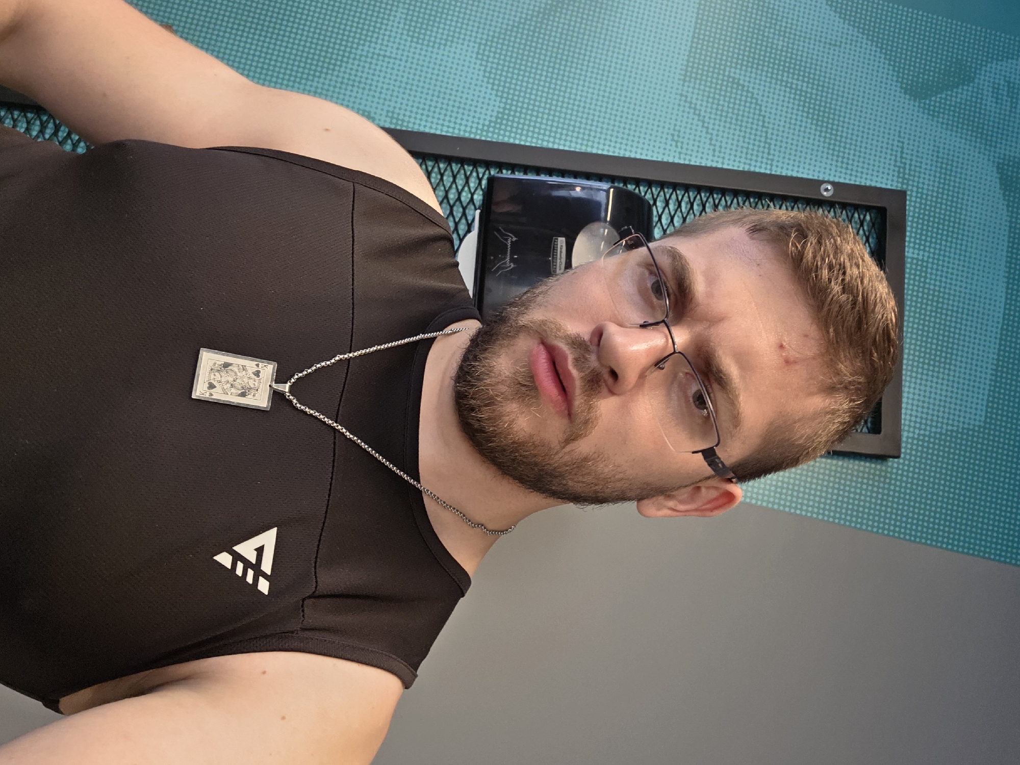 A close-up shot depicts a man wearing a black athletic tank top with a white triangular logo on the left side. He has short, light brown hair and a beard. He is wearing glasses with thin black frames. A silver chain necklace with a playing card pendant hangs around his neck. The man is looking directly at the camera with a somewhat neutral expression. Behind him is a metal grid structure with a black object inside. The background consists of a wall with a pattern of light blue dots on a darker blue base, transitioning to a solid gray wall. The location in Plovdiv, Bulgaria is indiscernible from the given visual information.