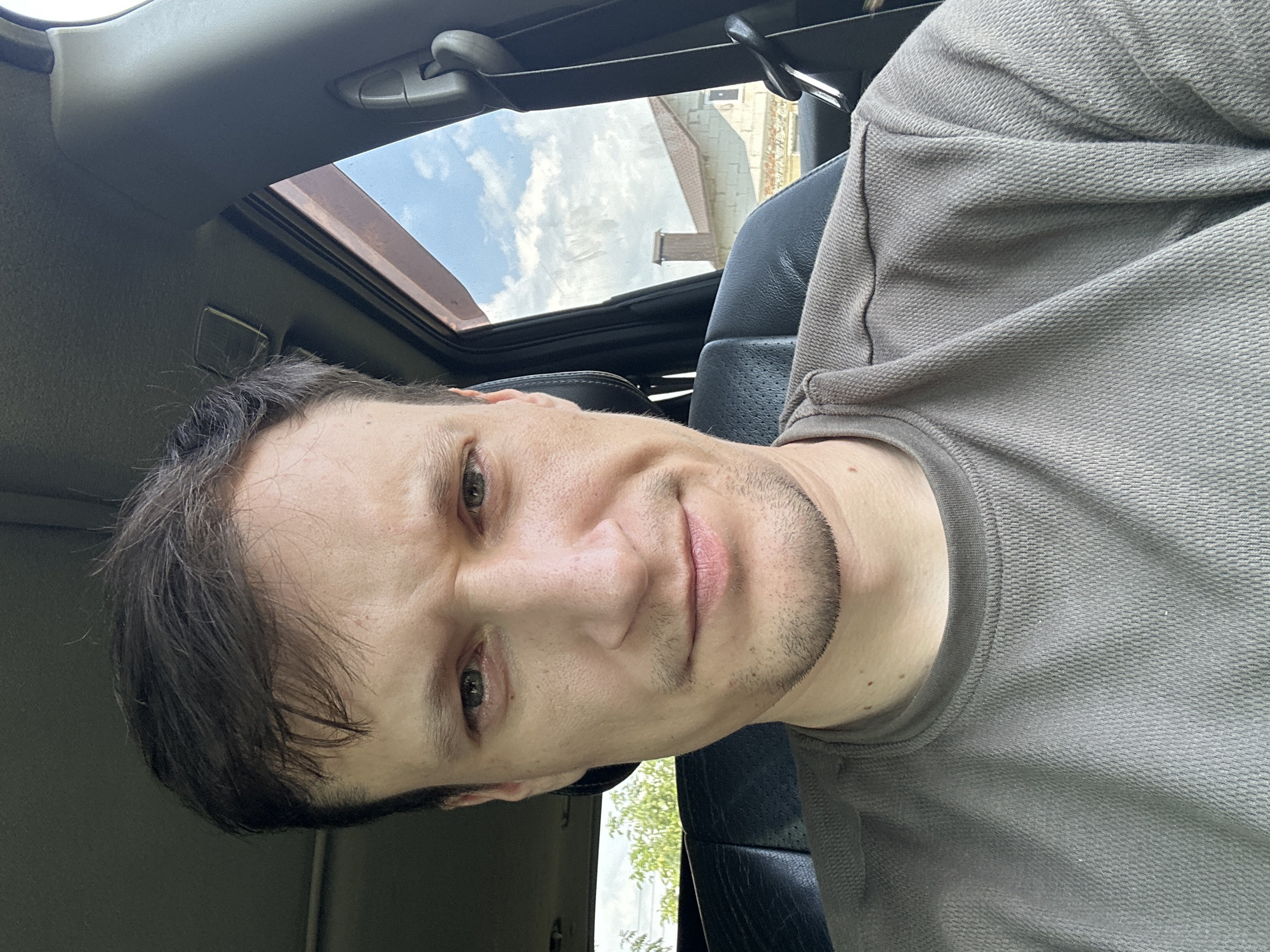 A man is sitting in the driver's seat of a car, looking at the camera. He has short, dark brown hair and is wearing a gray t-shirt. The car interior is visible, with the passenger seat and a window showing a residential area with houses and trees. The sky is blue with some clouds. The image appears to be taken on a sunny day.