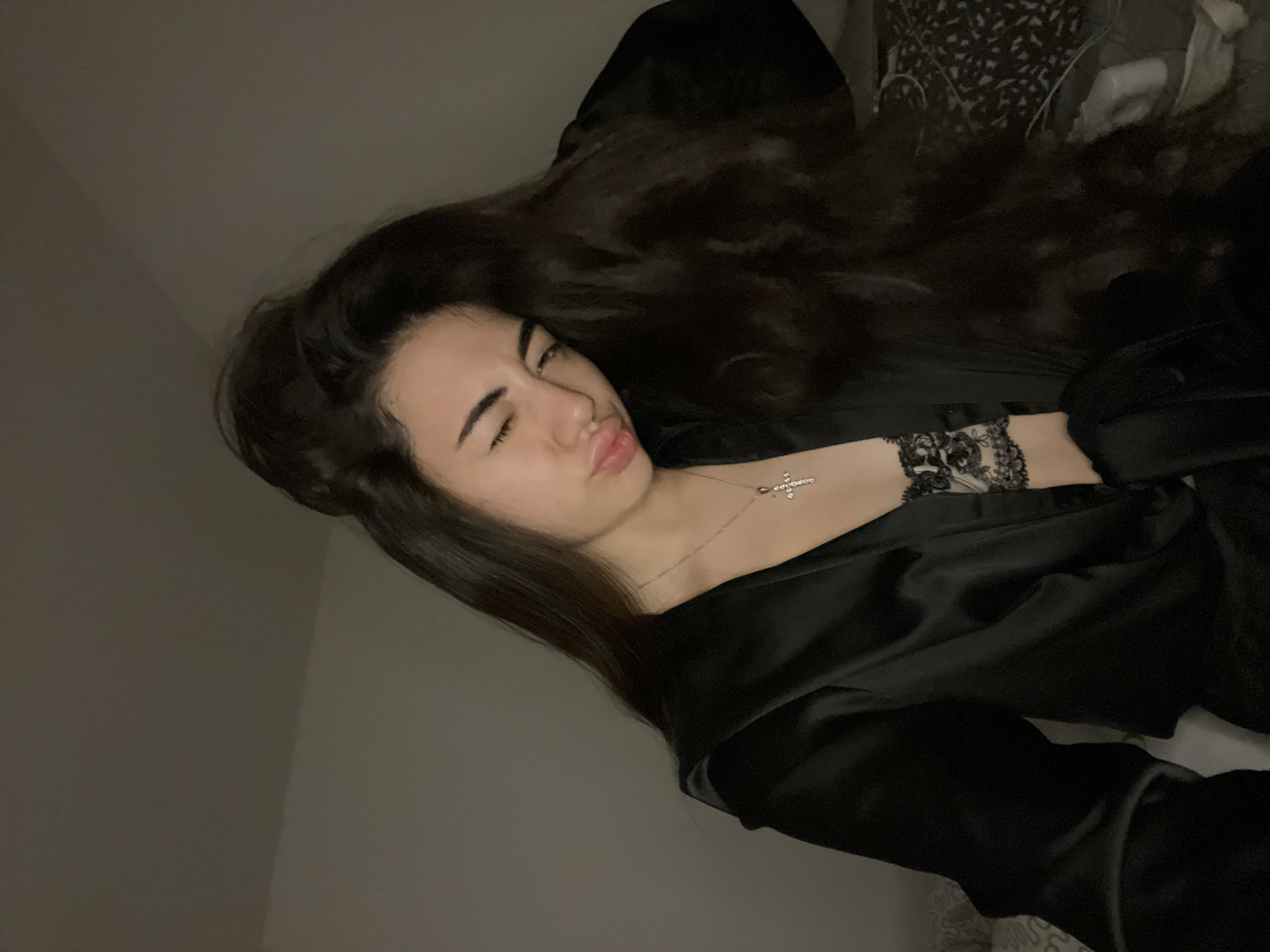 A young woman with long, dark, wavy hair is lying on her side in a dimly lit room. She is wearing a black satin robe over a black lace bra and a silver cross necklace. Her eyes are closed, and her lips are pursed. The background consists of a plain wall and a bed with patterned sheets. The room appears to be a bedroom. There is no indication of location within Chotěšov, Czechia.