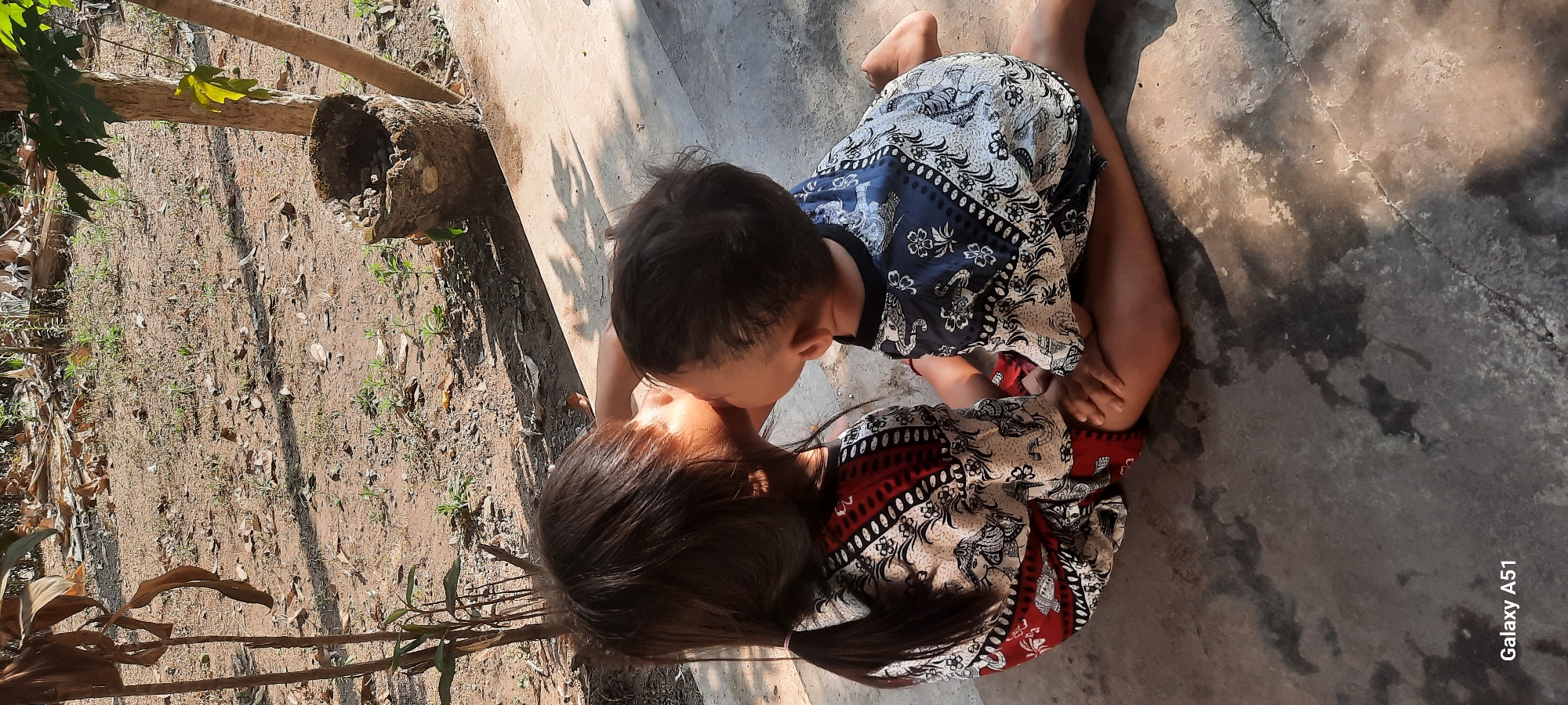 In Ploso, Indonesia, two young children are seated on a concrete surface, engaged in a tender moment. The older child, with dark hair tied in a ponytail, wears a patterned top in shades of red and white. They are leaning in, gently kissing the younger child's cheek. The younger child, facing away from the camera, has dark, short hair and is dressed in a blue and white patterned shirt. They are also seated, with their hands resting on their knees. The environment appears to be outdoors, with dry, dusty ground covered in sparse green shoots and fallen leaves. A rough, cylindrical object, possibly a woven pot or a hollowed-out log filled with small stones, sits near a tree trunk in the background. Sunlight casts dappled shadows on the ground and the children, suggesting a bright, sunny day. The lower right corner of the image displays "Galaxy A51".