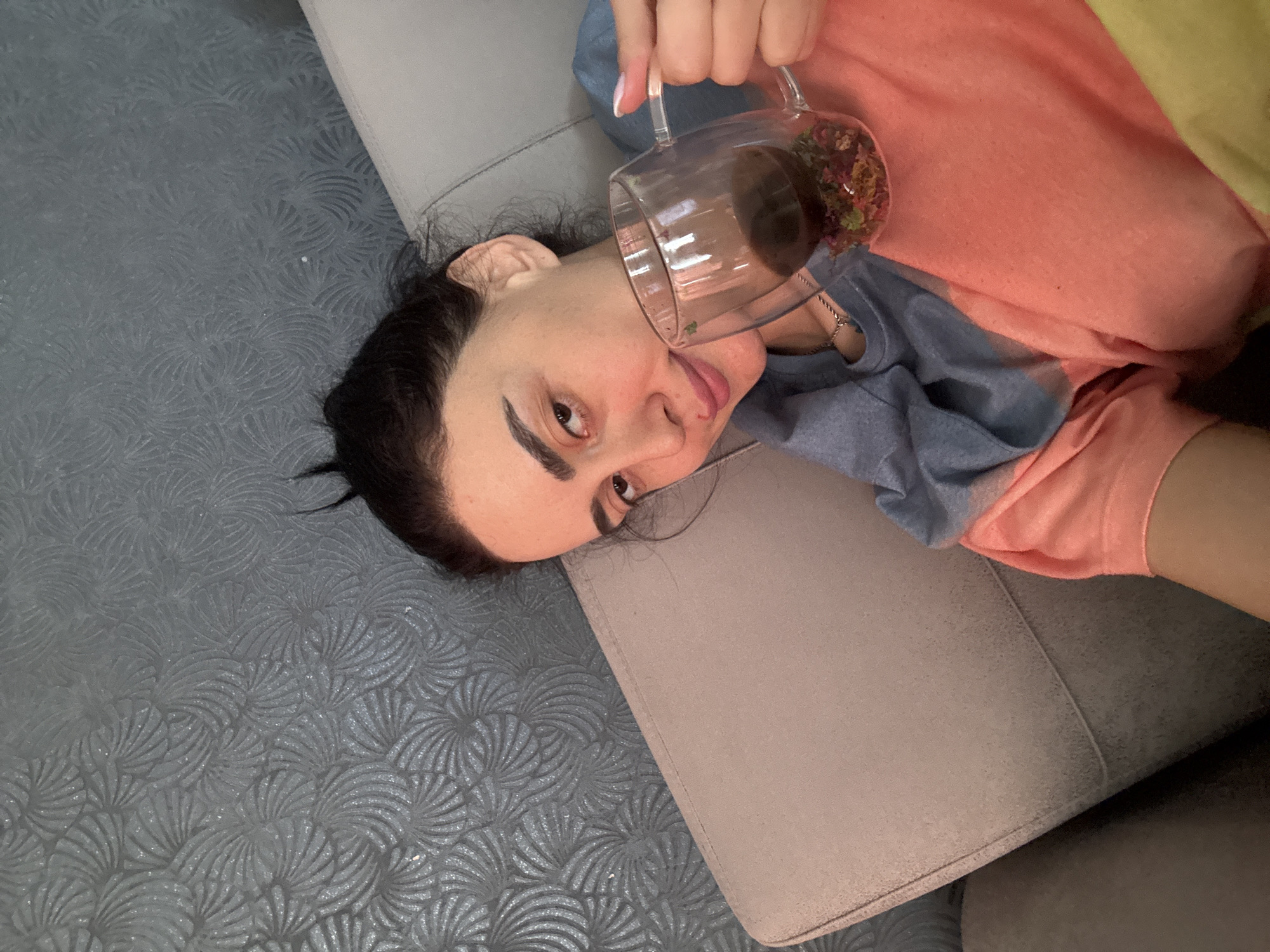 The image captures a playful indoor moment in what appears to be Tbilisi, Georgia. A person, likely female, with dark hair, prominent eyebrows, and brown eyes, is shown lying on their back on a light grey upholstered sofa. They are looking directly at the camera with a relaxed, slightly smirking expression, with their tongue playfully sticking out.
The individual is wearing a multi-colored top, primarily peach/coral and blue/grey, with a hint of yellow fabric visible in the background. In their right hand, they hold a clear glass mug with a handle, tilted towards the camera. The mug contains a dark liquid and a vibrant mixture of dried plant material, possibly flowers or herbs, at the bottom, suggesting a tea or decorative beverage. The person's fingernails are long and styled with a French tip.
The background features a dark grey floor or carpet with a repeating, textured fan-like or radial pattern. The overall atmosphere conveyed is one of casual relaxation and lightheartedness. The time of day is not discernible, and no weather conditions are visible from this indoor setting. There are no specific visual cues within the image itself to confirm the Tbilisi, Georgia location. No text is visible in the image.