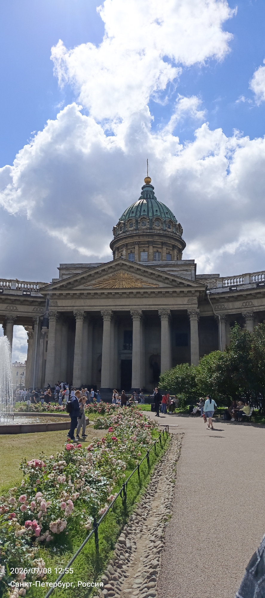 This is a photograph of Kazan Cathedral in Saint Petersburg, Russia, on a partly cloudy day. The cathedral's large green dome and classical facade with Corinthian columns dominate the background. In the foreground, a pathway curves through a garden filled with blooming pink and white roses. Several people are visible in the scene: some are walking along the pathway, others are sitting on benches or standing near the garden, and a group is gathered near the entrance of the cathedral. A fountain is visible to the left of the frame. The timestamp and location "2026/07/08 12:55 Санкт-Петербург, Россия" are superimposed on the bottom left of the image.