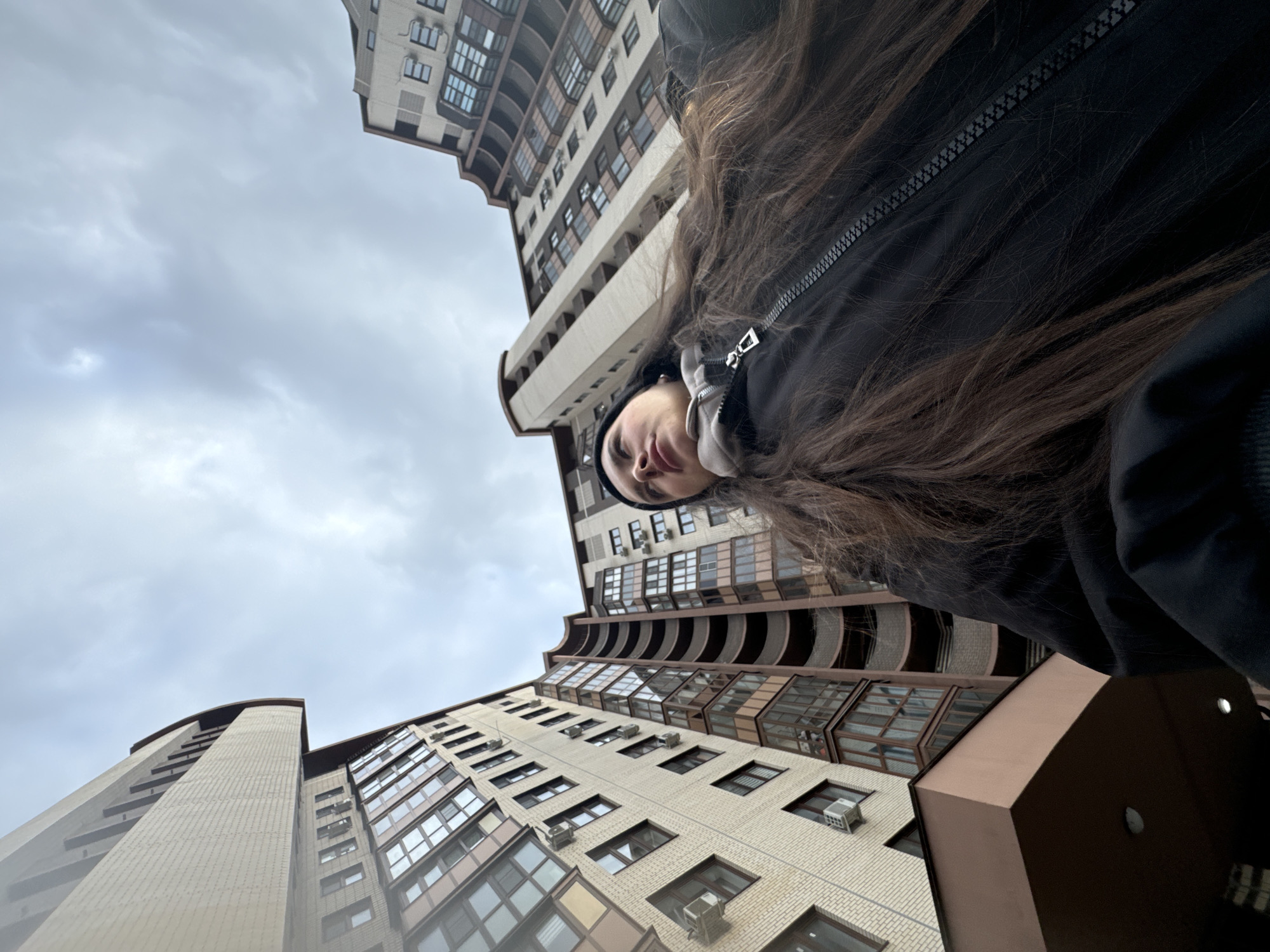 The image captures a unique low-angle perspective, looking upwards towards tall residential buildings and an overcast sky. In the foreground, positioned at the top of the frame, is a person, likely female, with long, dark brown hair cascading downwards. She is wearing a black zippered jacket and possibly a dark beanie or hood, with only the upper part of her face visible as she looks down towards the camera with a neutral expression.
Two prominent, modern apartment buildings frame the scene. They are multi-story structures with light-colored brick or facade, featuring numerous windows and enclosed balconies with darker frames. Several air conditioning units are visible on the exterior walls of the buildings. The architecture suggests a slight curve to the buildings' design. The sky is uniformly grey and cloudy, indicating an overcast day with no direct sunlight. The scene depicts an urban or suburban environment in Akulovo (Gp Kubinka), Russia. The activity appears to be a self-portrait taken from a low viewpoint, looking up at the subject and the surrounding architecture. No visible text is present in the image.