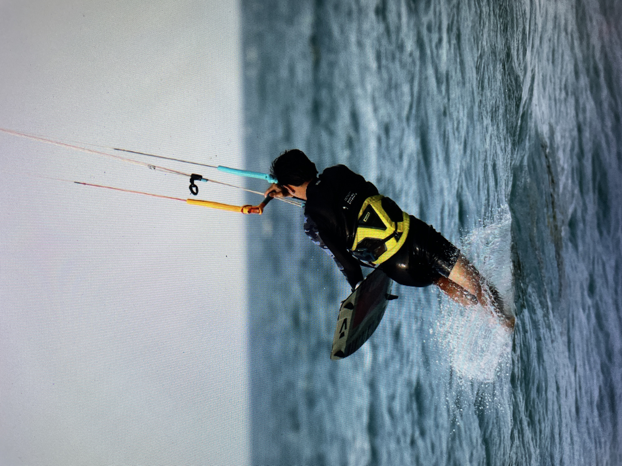 A person is kitesurfing on the ocean. The person is wearing a black wetsuit with a yellow safety vest and is holding onto the kite with a bar that is colored blue and yellow. A kiteboard is attached to the person's feet. The water is choppy, and there are waves. The sky is overcast. The location is Bacong, Philippines.